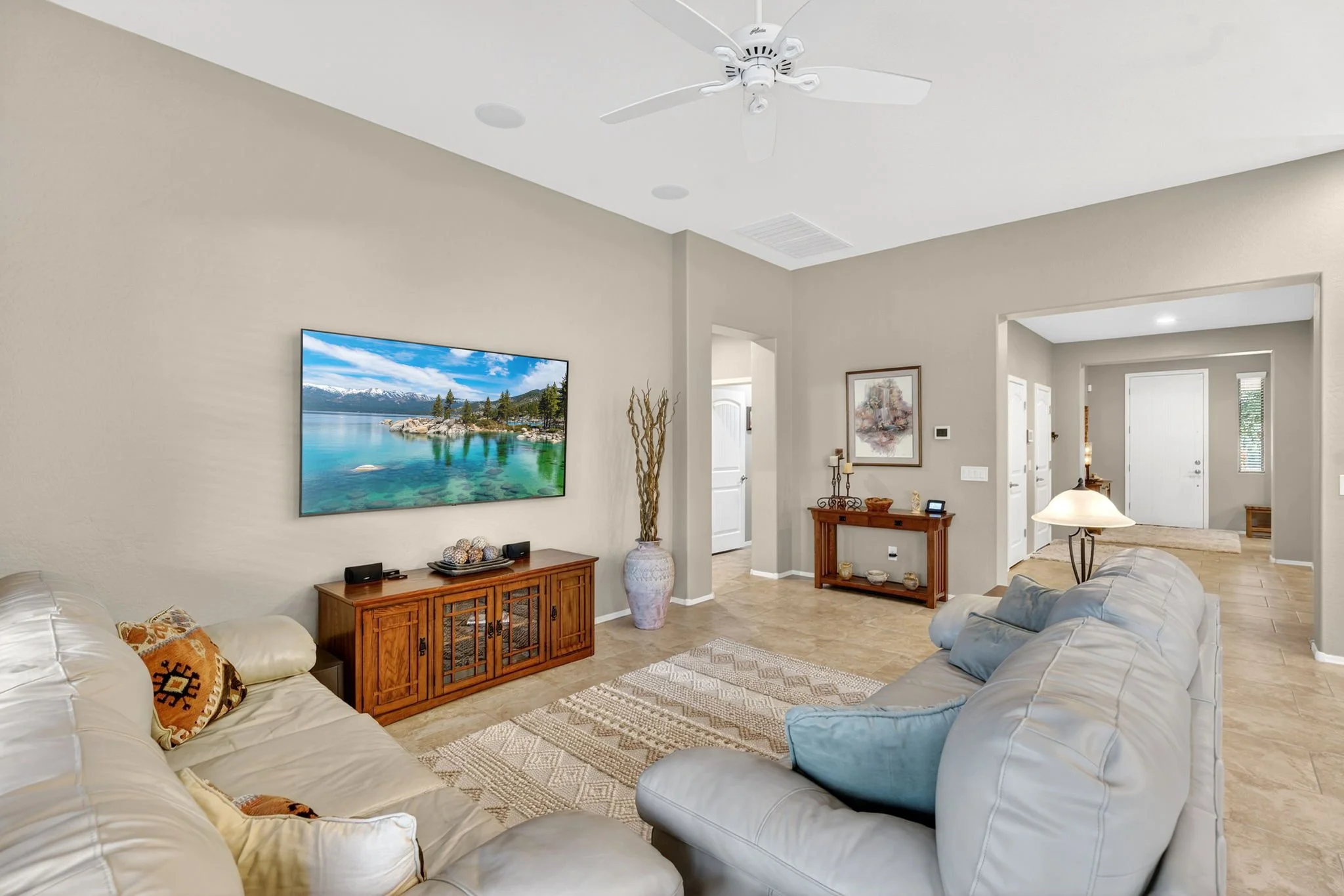 Living room with beige walls and two white sofas, a TV mounted on the wall showing nature scene, wooden furniture, and a chandelier hanging from the ceiling.