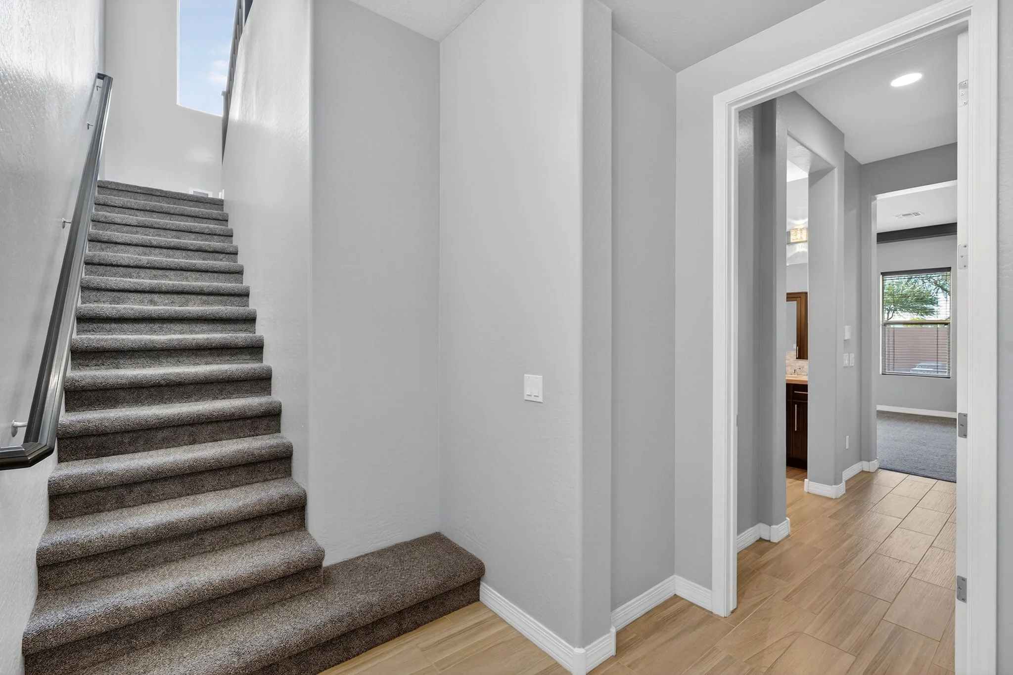 Interior of a modern house showing a staircase with gray carpet, white walls, and a hallway with wooden flooring leading to other rooms.