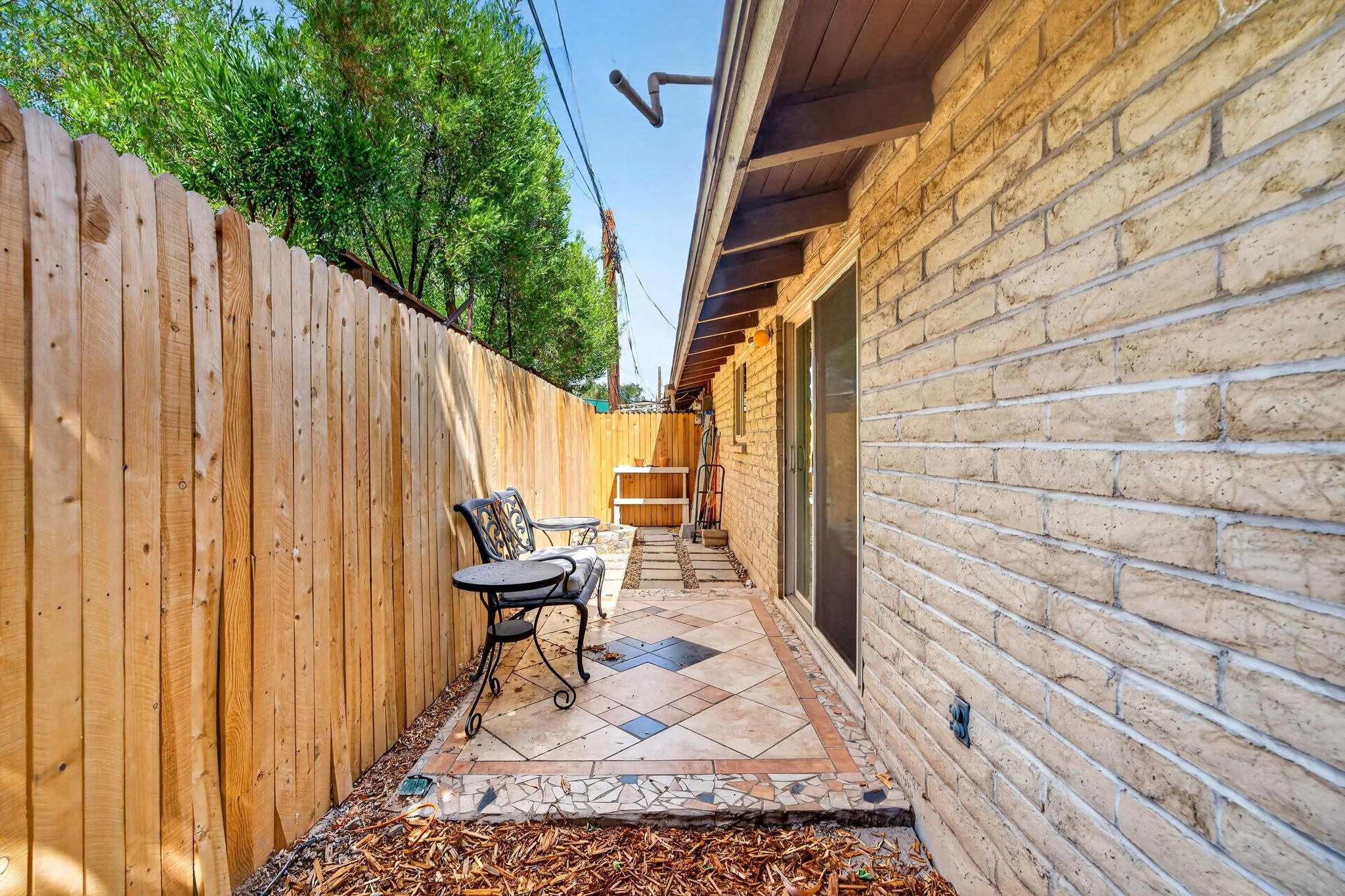 A narrow backyard patio with a bench, two small tables, and a storage box against a brick wall, enclosed by a wooden fence, with trees and a blue sky in the background.