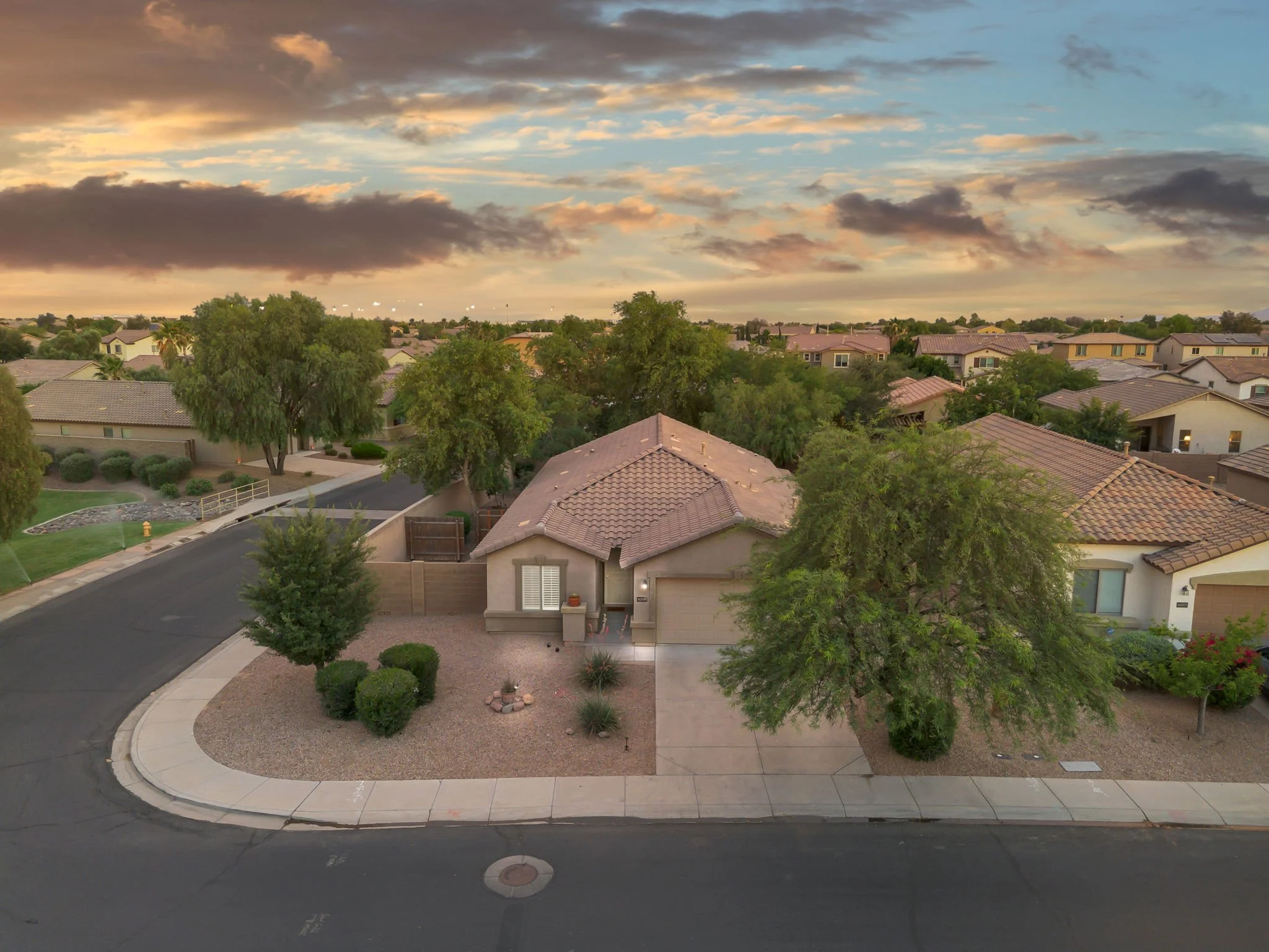 A suburban neighborhood with single-family homes, trees, landscaping, and a street at sunset with a partly cloudy sky.