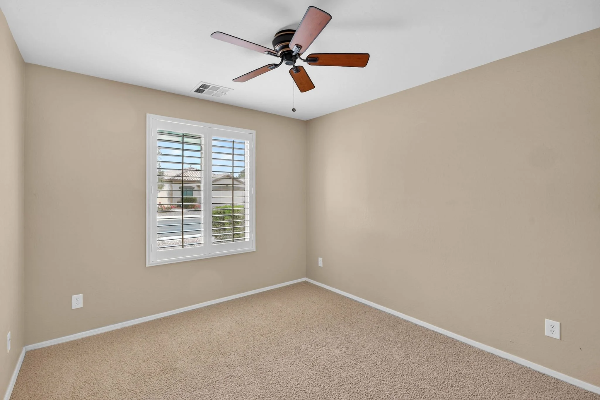 Empty beige-colored room with a window that has white shutters, a ceiling fan with wooden blades, carpeted flooring, and electrical outlets on the walls.