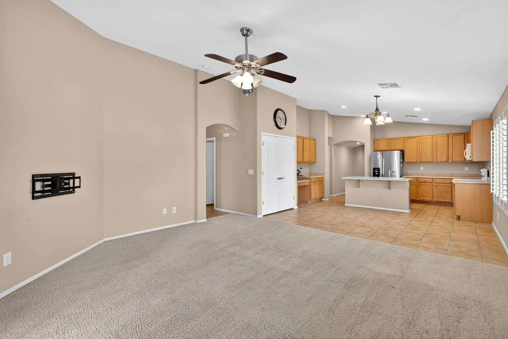 Open-concept living space with beige walls, light-colored carpet, kitchen with wooden cabinets, stainless steel refrigerator, tiled floor, ceiling fan, and multiple light fixtures.