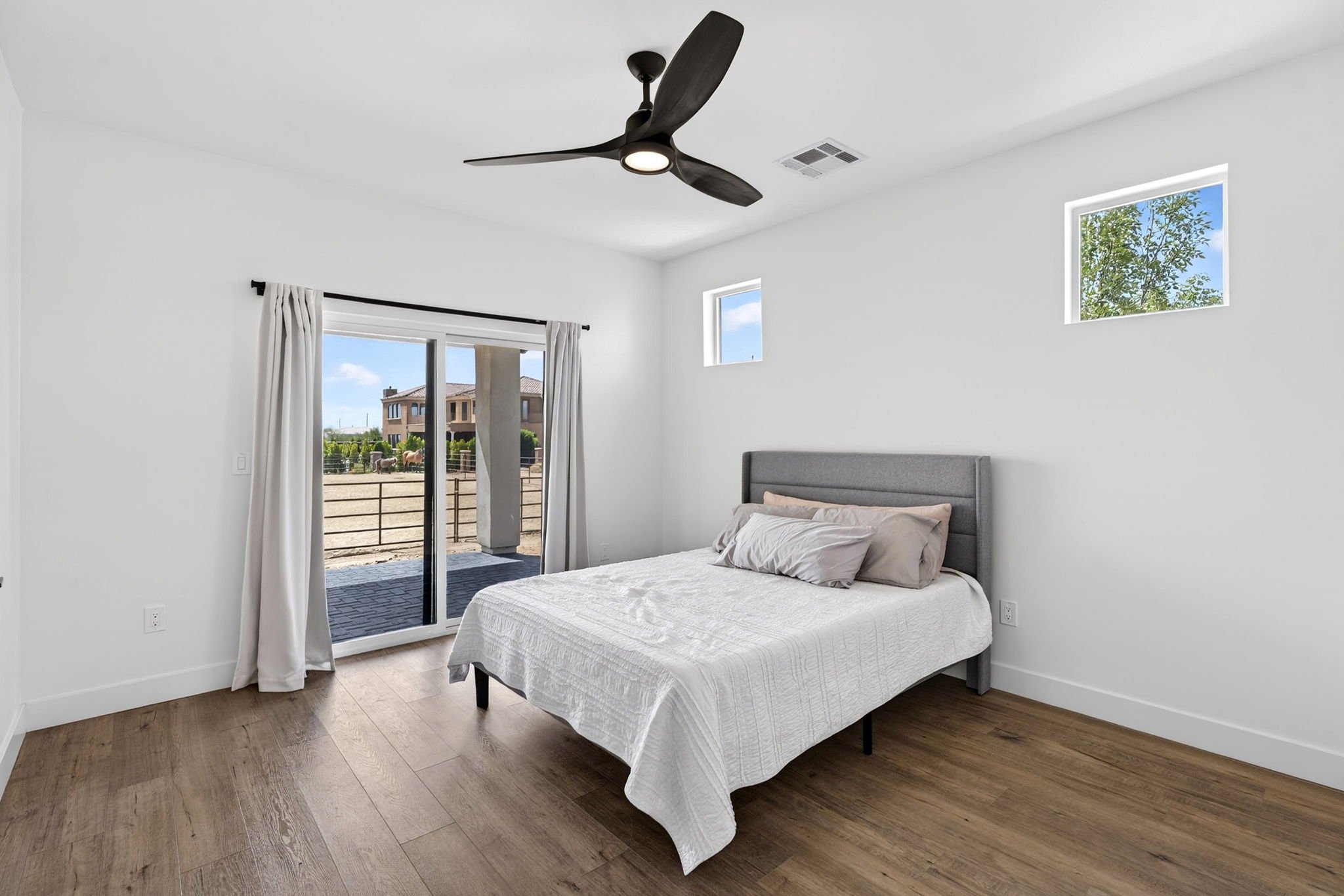 A modern bedroom with white walls, wooden flooring, a bed with a gray headboard and white bedding, two small rectangular windows, and a sliding glass door with white curtains leading to an outdoor area with a fence and neighboring houses. A black cei