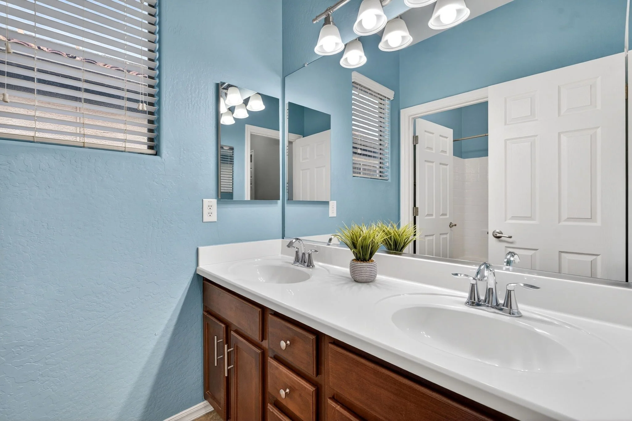 A bathroom with a white countertop, dark wood cabinet, and large mirror. Blue textured walls, a window with blinds, ceiling lights, and a door.