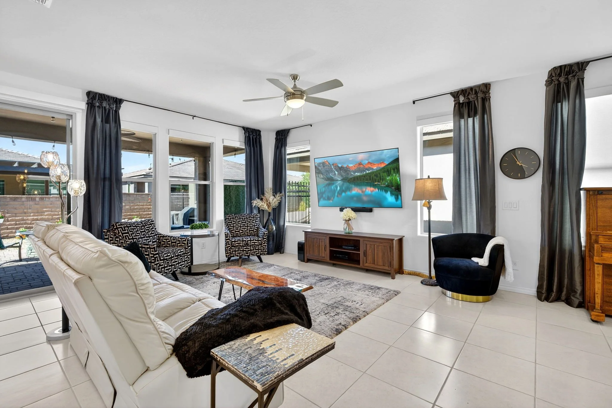 Living room with white walls, large windows with dark curtains, a ceiling fan, a flat-screen TV, various chairs and sofas, a wooden TV stand, a floor lamp, and decorative items such as flowers and a clock.