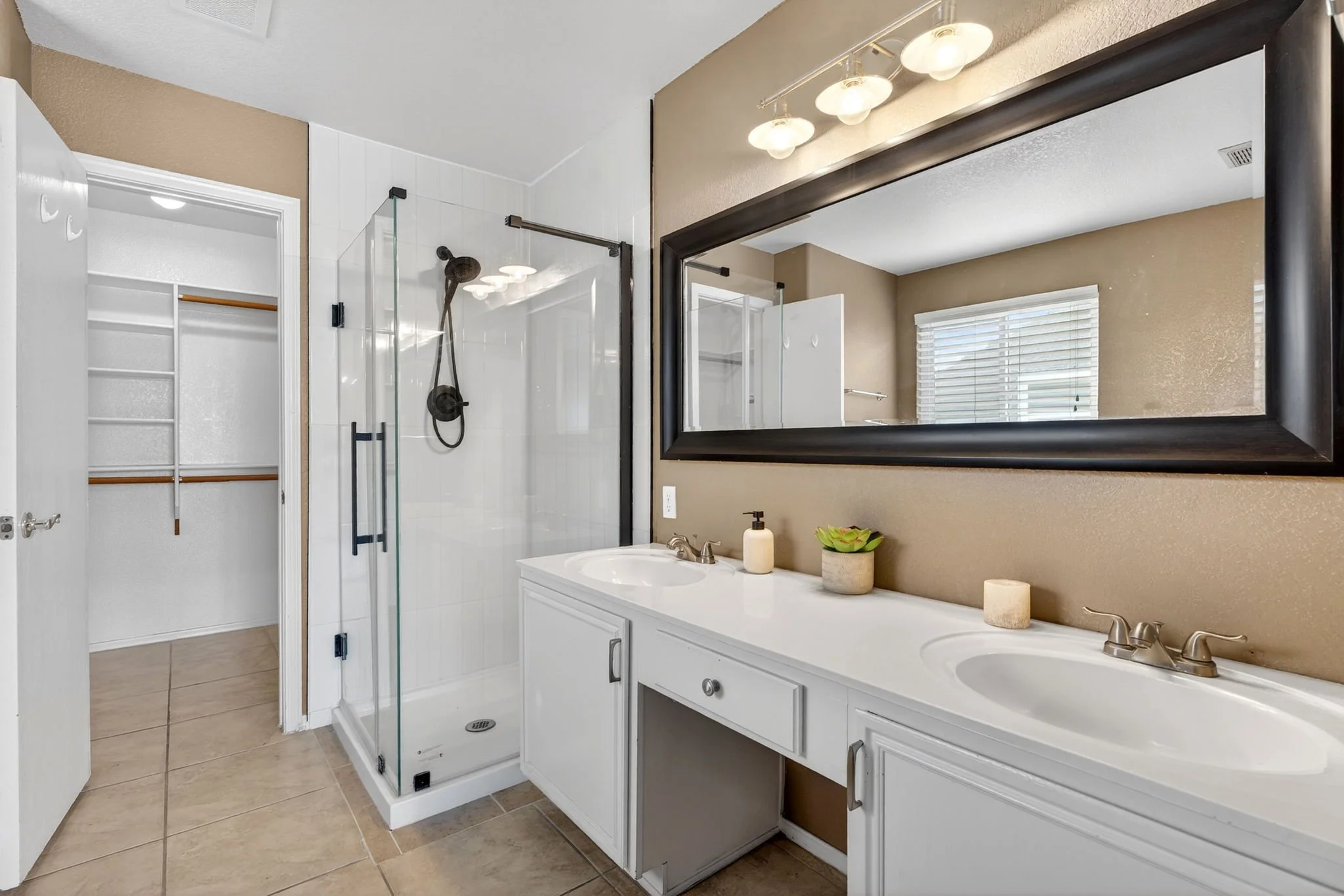 A bathroom with a double sink vanity, large mirror, and three light fixtures, a glass-enclosed shower with a black showerhead, a built-in linen closet, and a window with blinds.