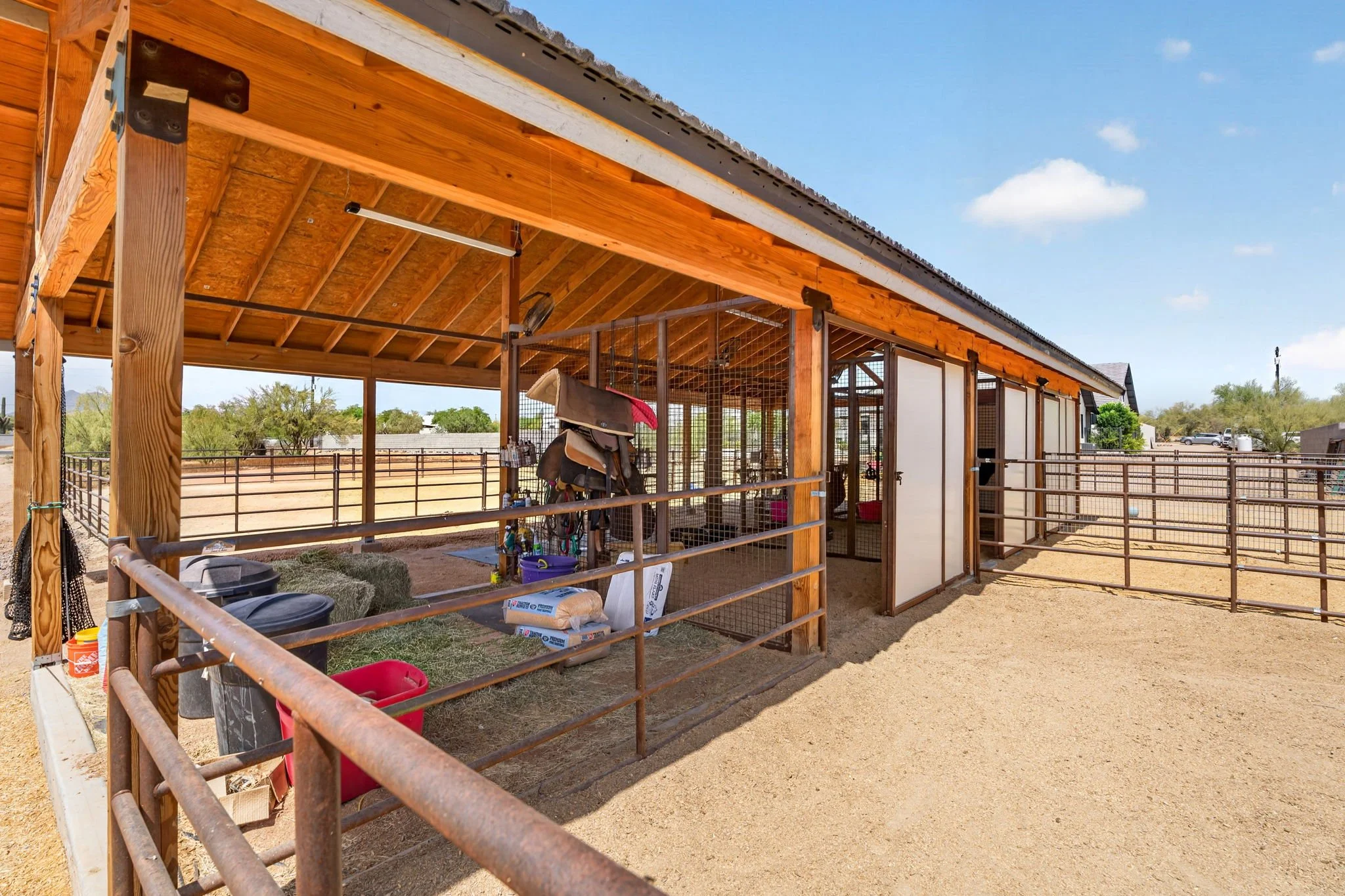 Wooden horse stable with a partially open roof, fencing, and various equipment inside, set in an outdoor rural area with clear blue sky and some trees in the background.