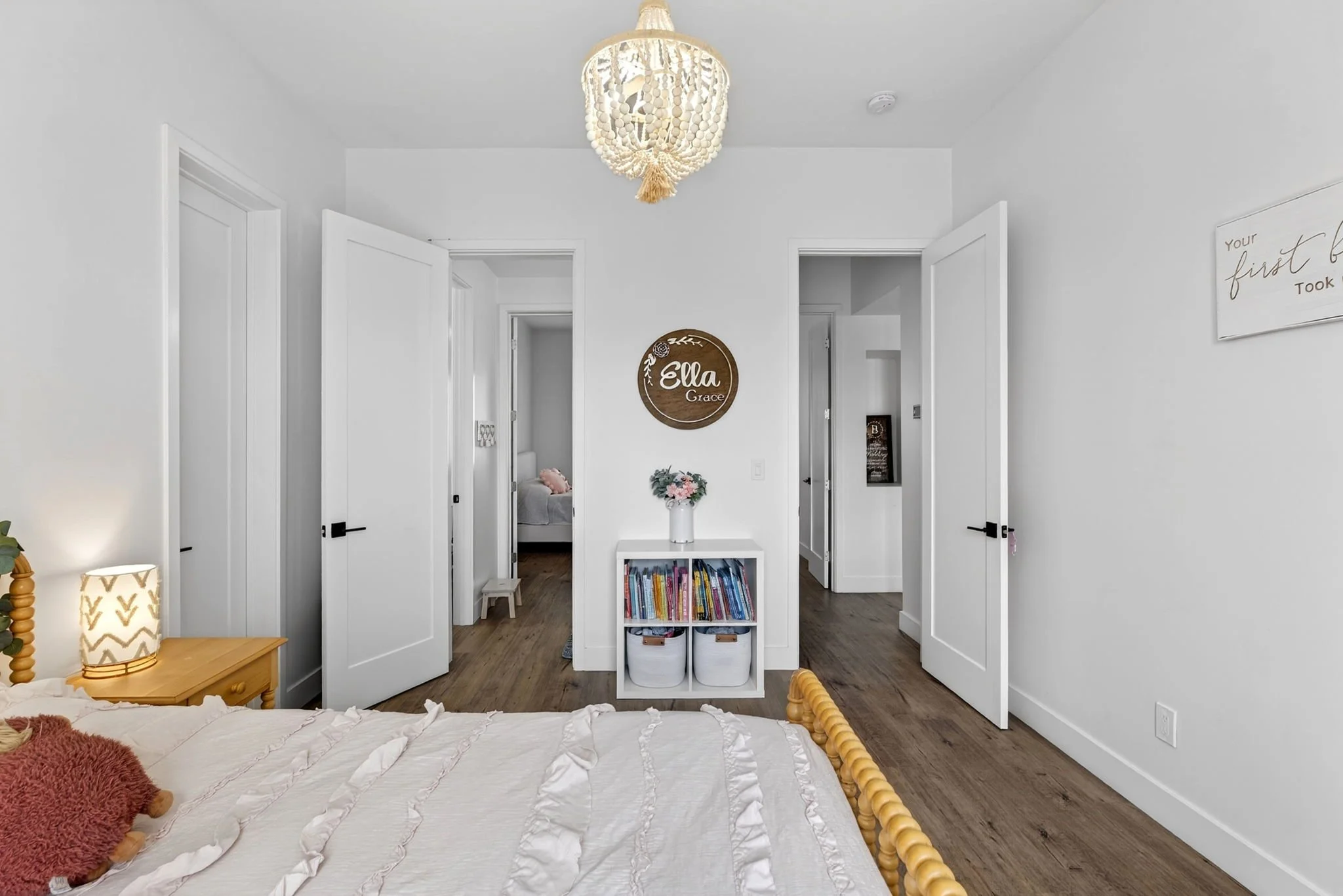 A bedroom with a white bed and a wooden nightstand with a patterned lamp. The room has white walls and doors, with a small bookcase at the center holding books and storage baskets. A chandelier hangs from the ceiling, and a wall sign that says 'Ella 