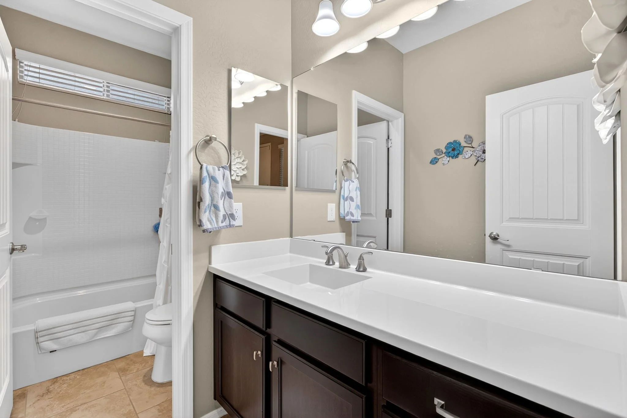 Bathroom with a shower, toilet, white vanity with dark cabinets, mirrors, and decorative wall art, beige walls, window blinds, and ceiling lights.