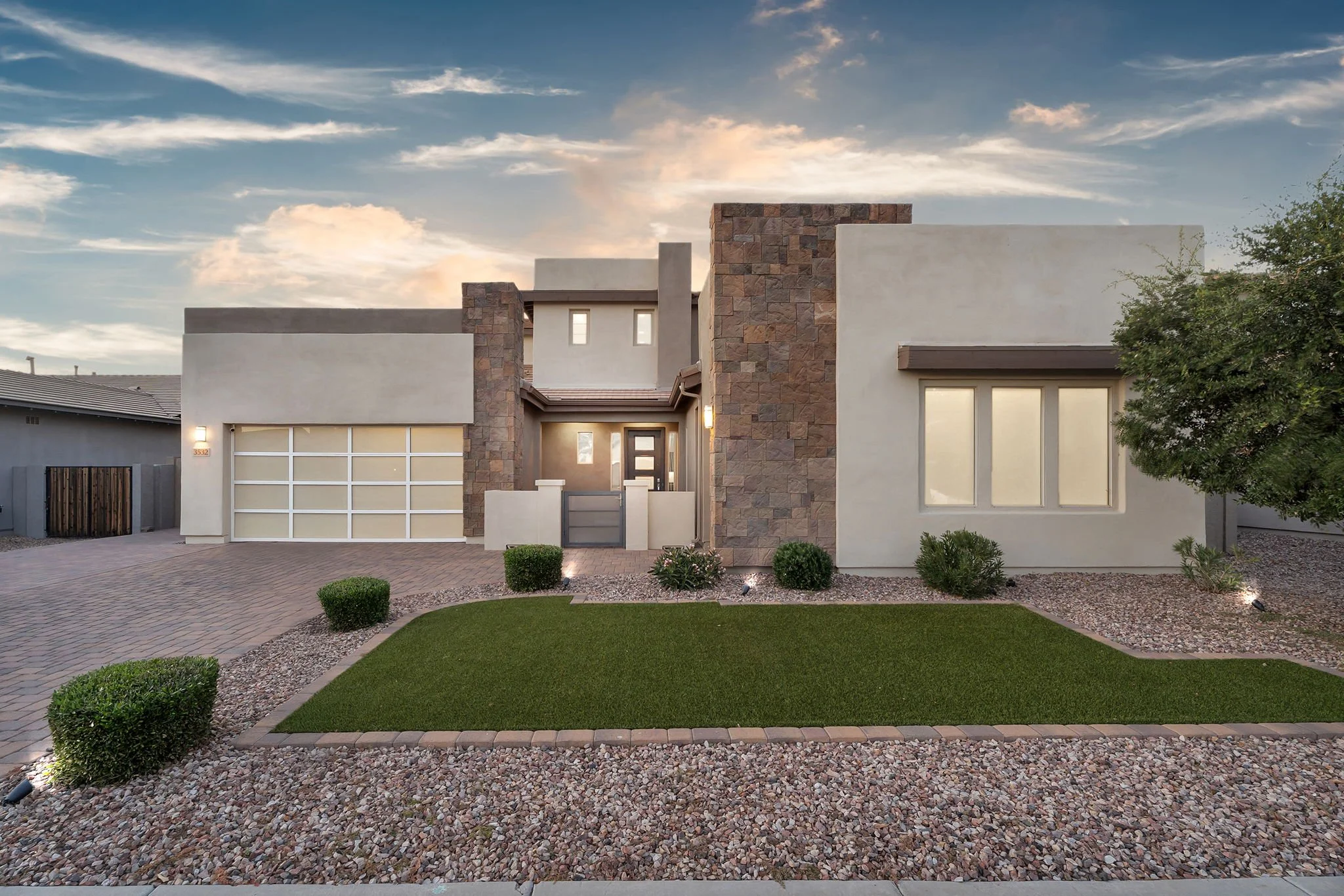 Modern house with white exterior walls, stone accents, large windows, a garage with a white door, and a well-maintained front yard with small bushes and gravel landscaping.