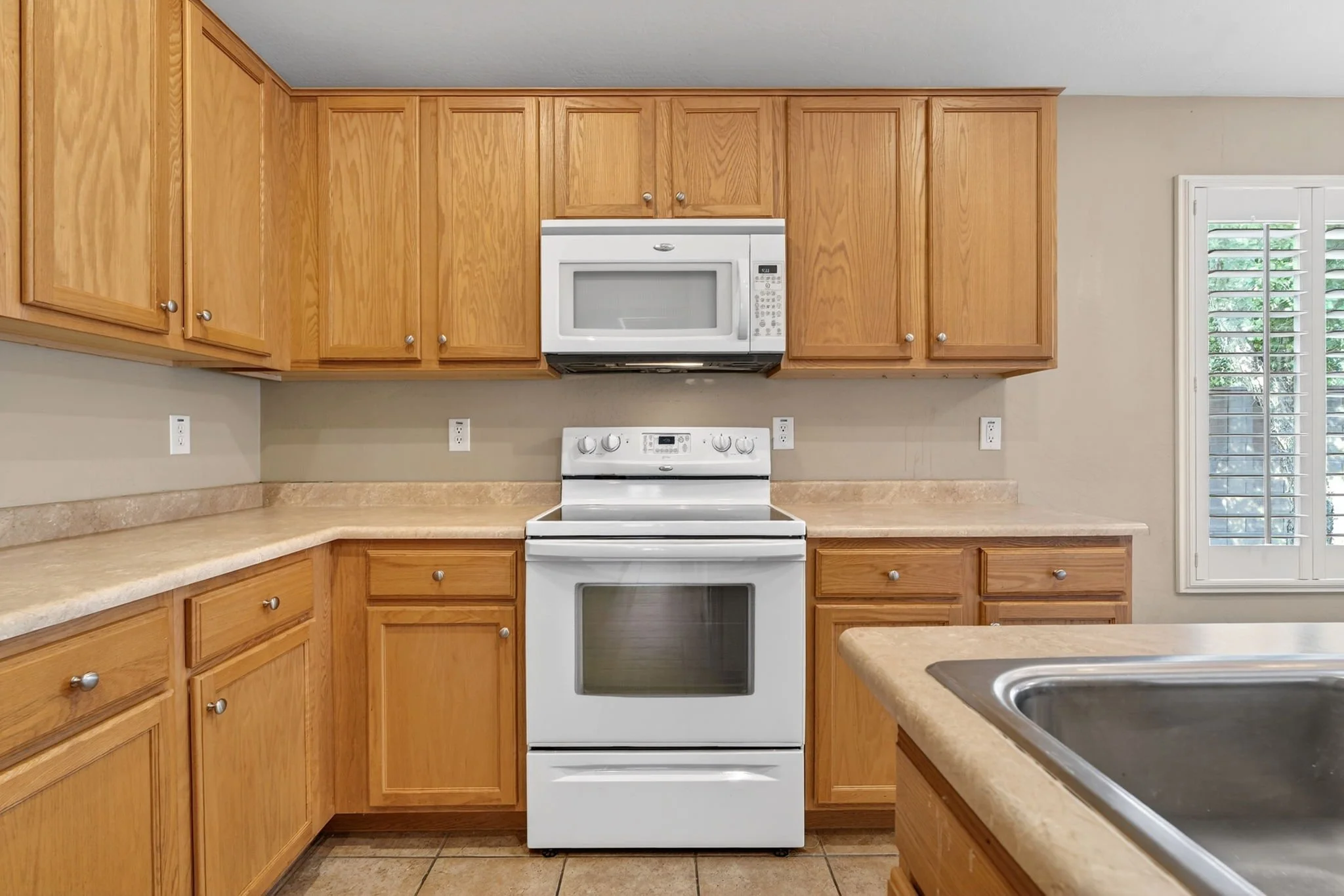 Kitchen with wooden cabinets, white stove and microwave, beige countertop, and a window with white shutters.
