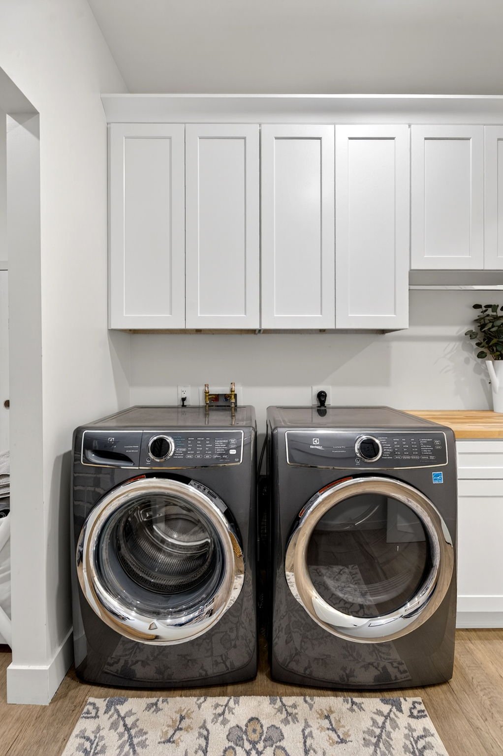 Front view of a laundry room with white cabinets above a black washer and dryer, a wooden countertop on the right, and a potted plant.