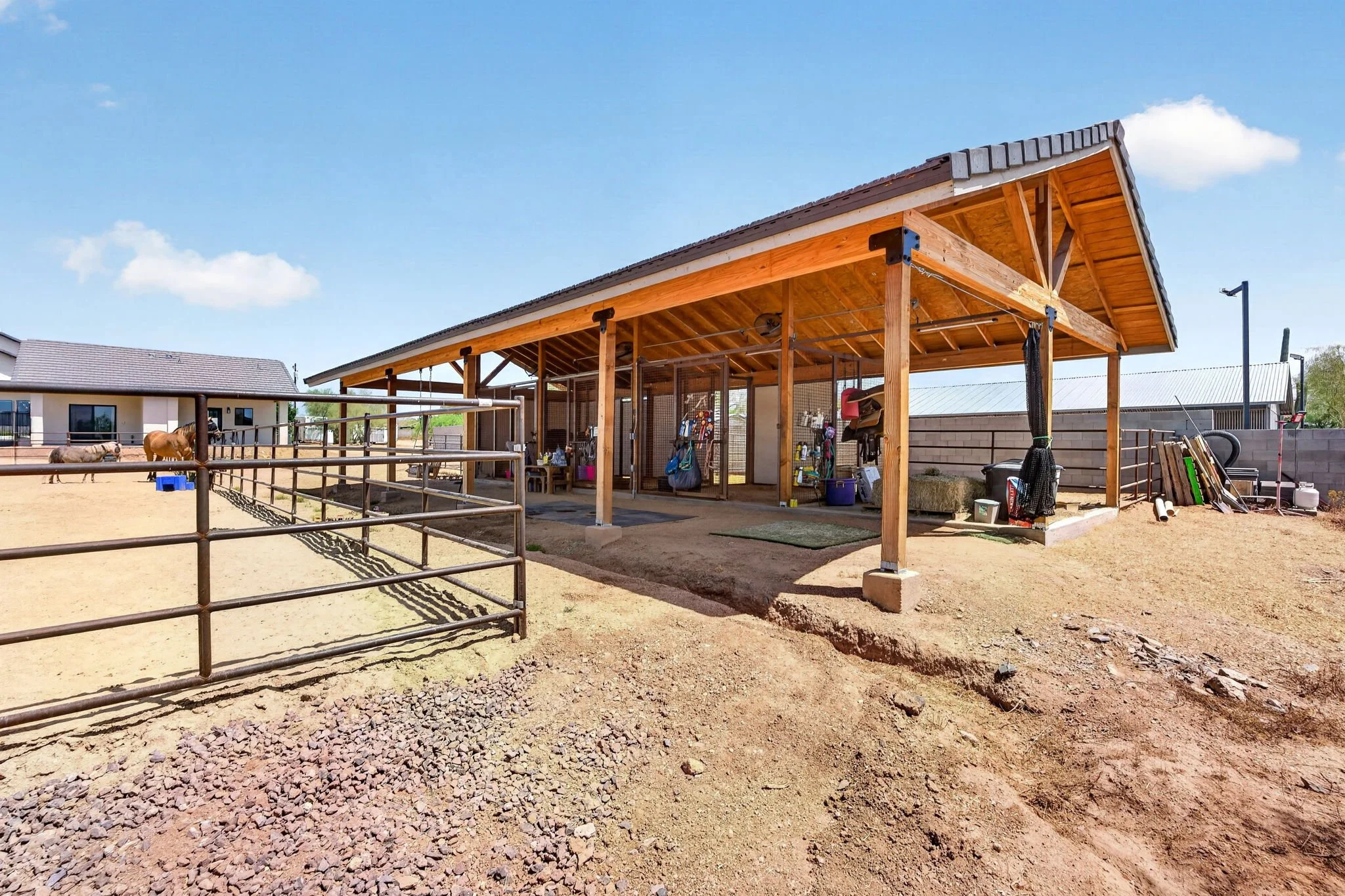 A wooden shelter at a horse stable with a fenced area, horses, and equestrian equipment under a clear blue sky.