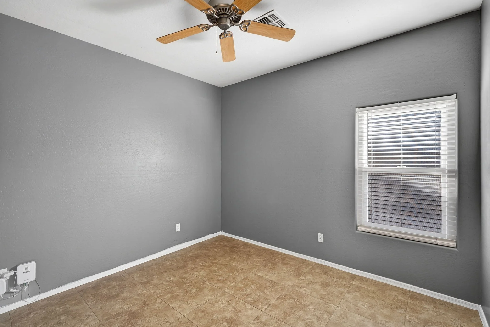 Empty room with gray walls, a window with blinds, a ceiling fan with wooden blades, and tiled flooring.