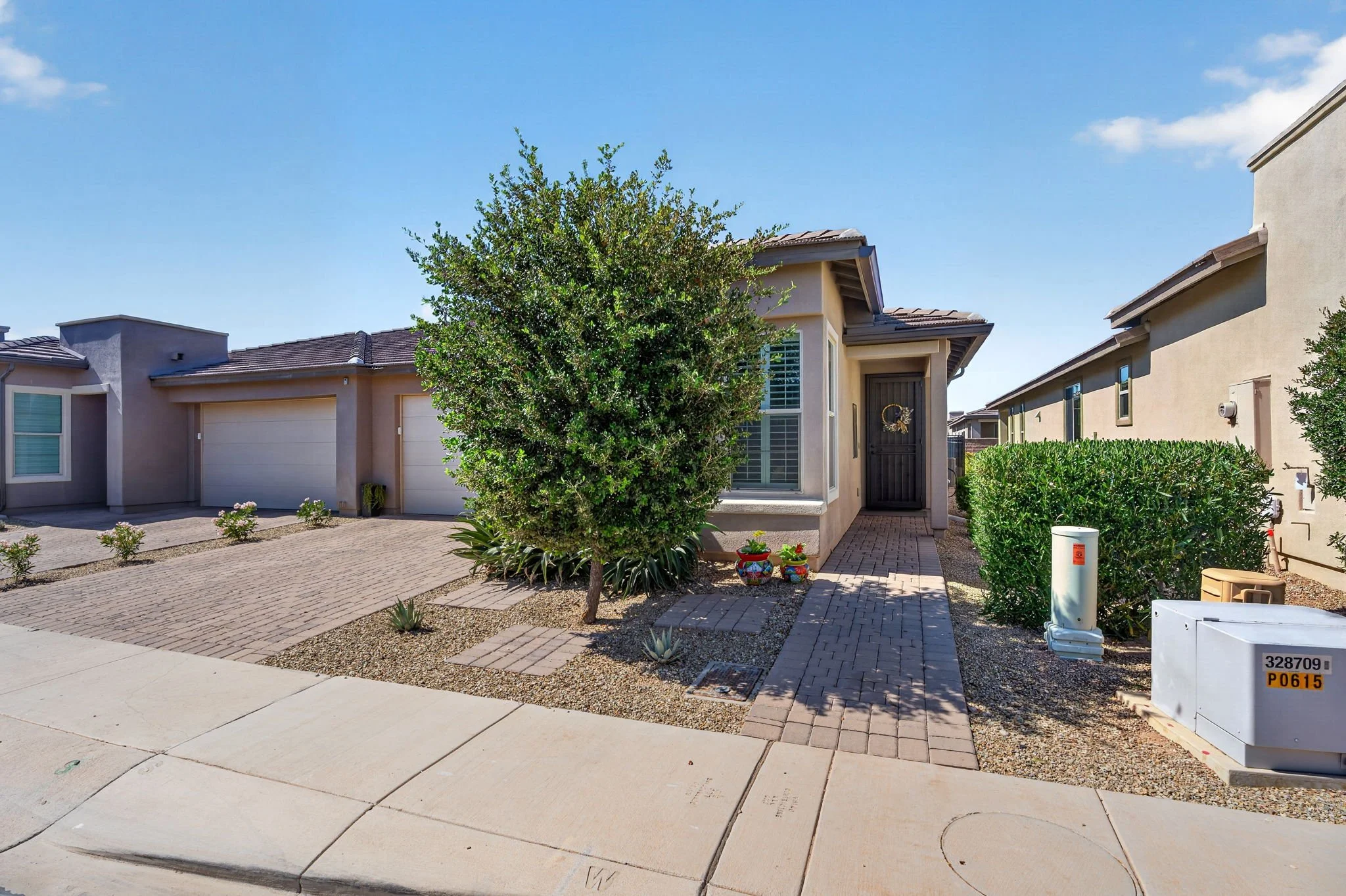 Front yard of a modern beige house with a tree, small bushes, and decorative pots. Paved walkway and driveway with neighborhood houses in the background.