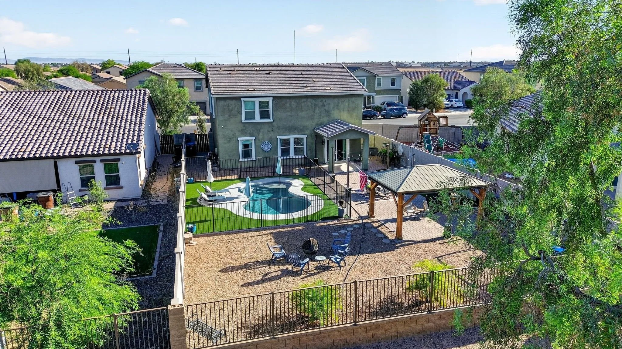 A backyard with a small kidney-shaped swimming pool, surrounded by a black metal fence, with lounge chairs and umbrellas. There is a wooden pergola with seating and a small paved area. Neighboring houses with tile roofs are visible in the background 