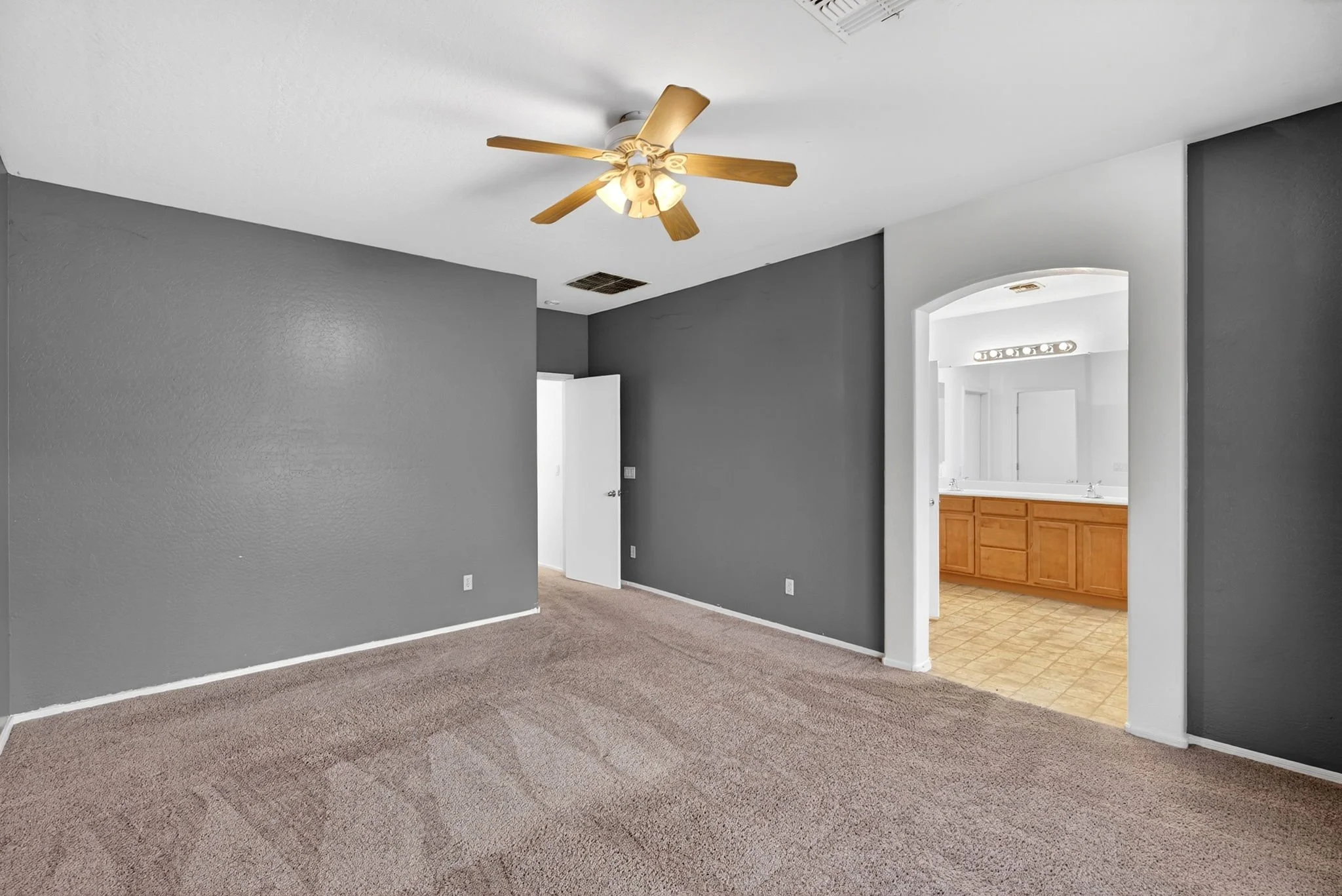 Interior of a house with gray walls, a ceiling fan, beige carpet, an arched doorway, and a view into a kitchen with wooden cabinets and a tiled floor.