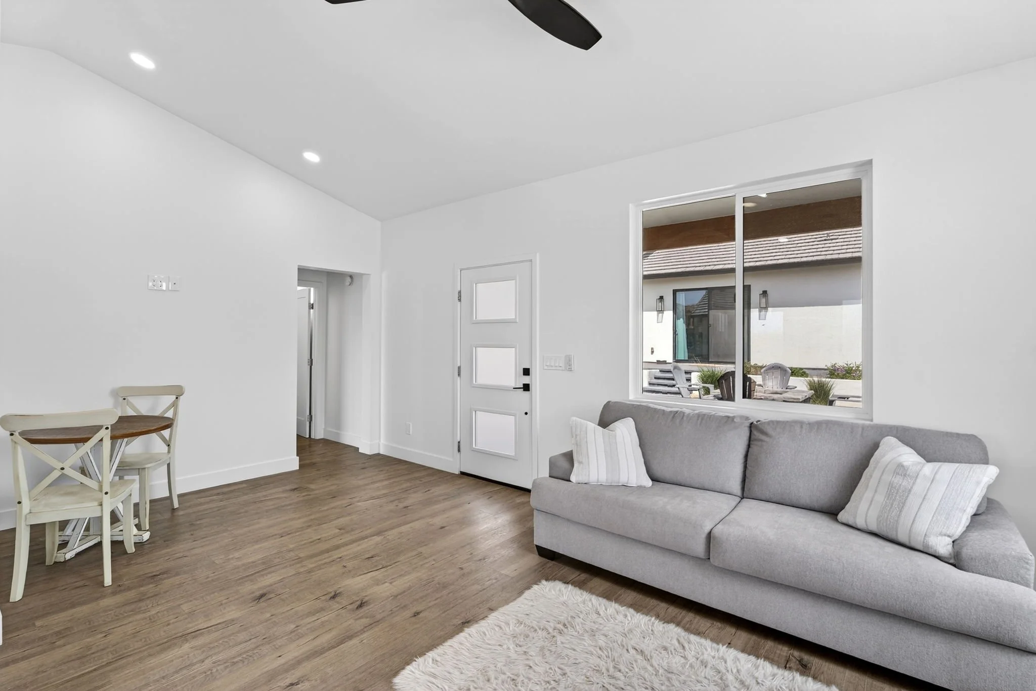 A living room with white walls, a grey couch with striped pillows, a white shag rug, wooden floor, a window with outdoor patio view, and a small dining area with a round table and four chairs.