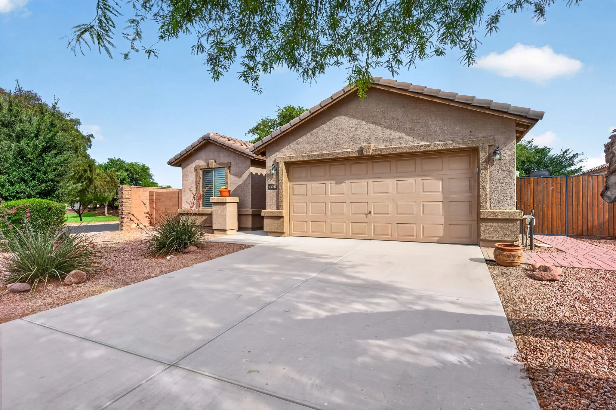 A beige stucco house with a two-car garage, desert landscaping, and a concrete driveway, under a partly cloudy sky.