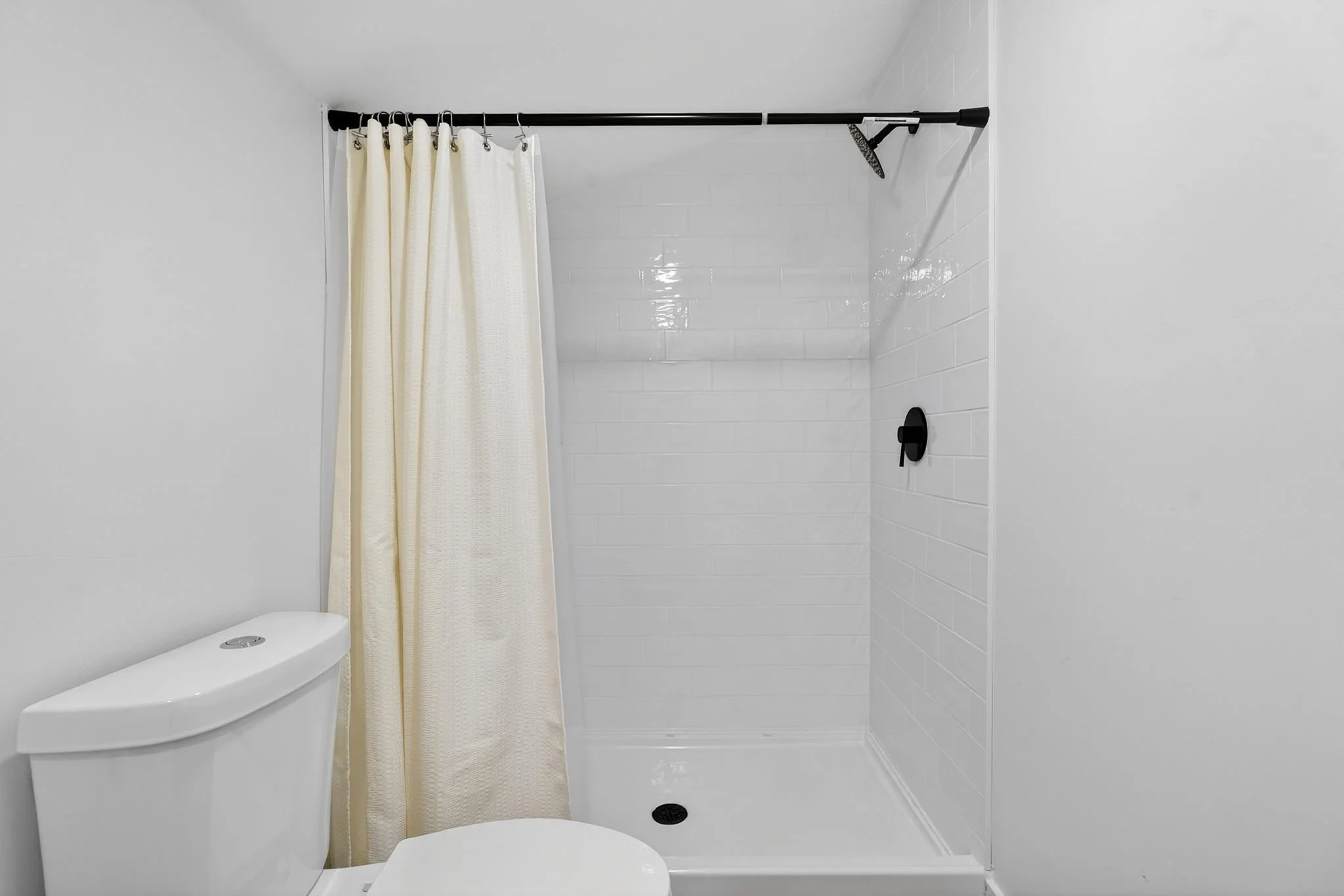 A modern, minimalistic bathroom with white walls and white tiled shower area. A black showerhead is mounted on the wall, with a cream-colored shower curtain hanging on a black rod. There is a white toilet in the foreground.