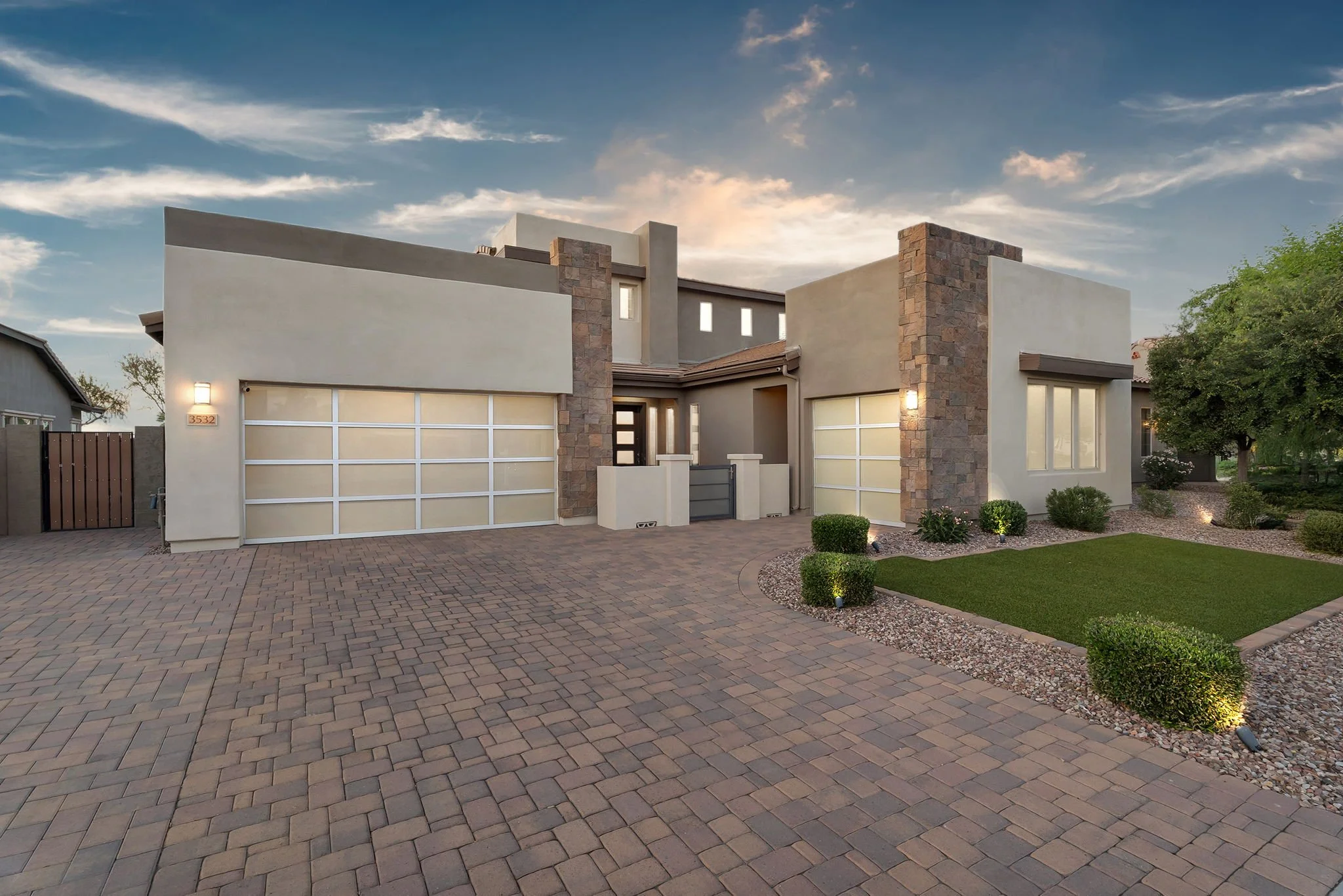 Modern two-story house with a beige exterior, stone accents, large windows, garage, well-maintained front yard with small bushes, and a brick driveway under a sky with scattered clouds.