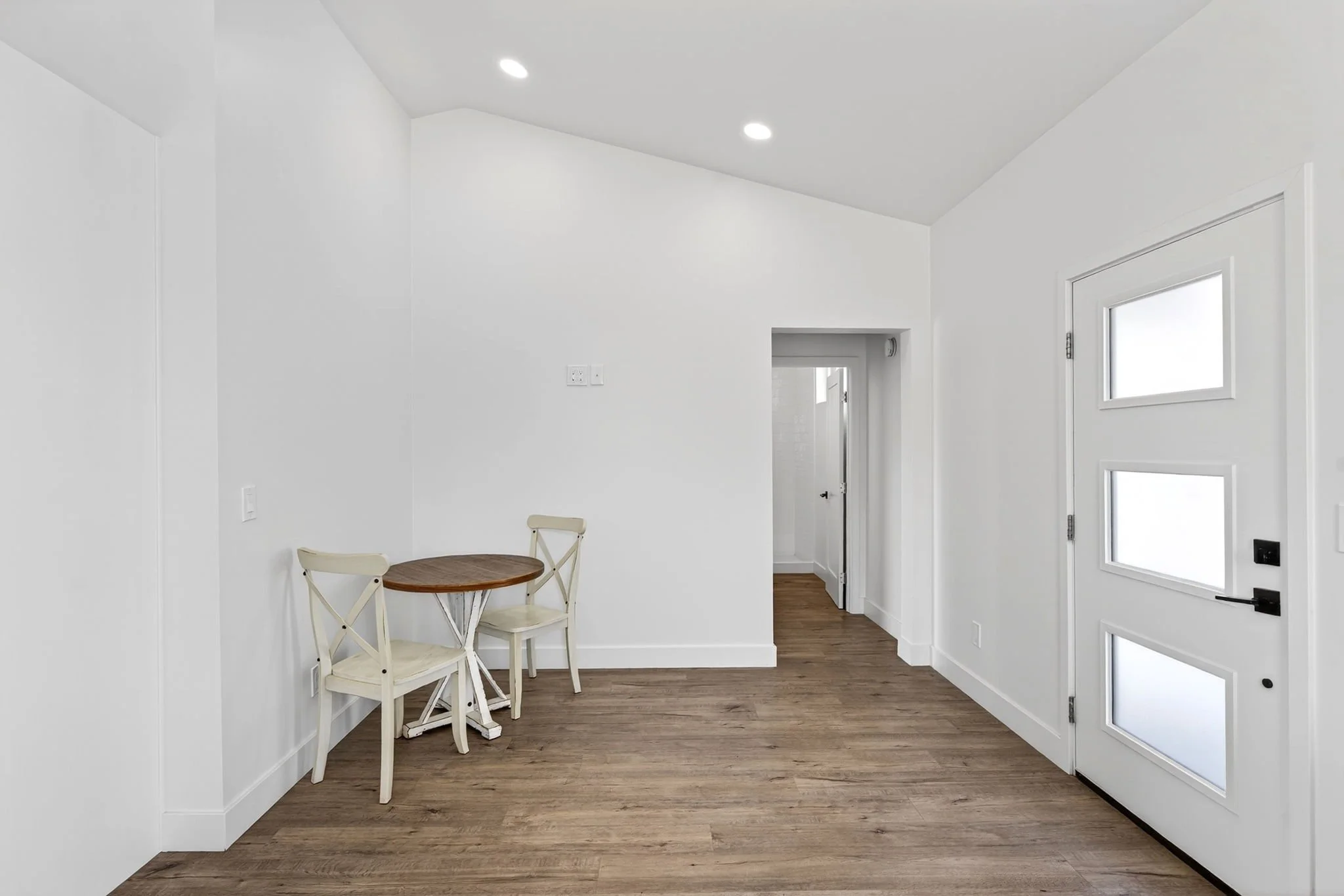 Interior view of a white modern room with a small round wooden table, two white chairs, a door with windows, wood flooring, and minimal decor.