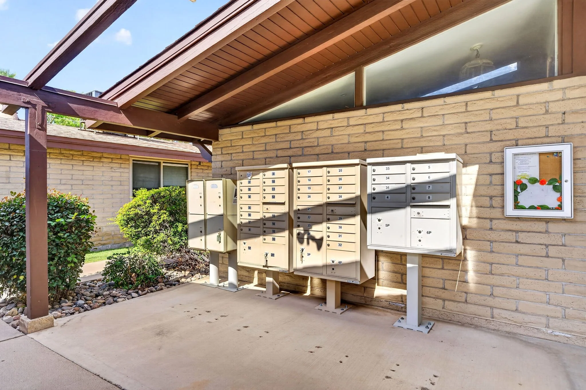 Apartment building entrance with multiple mailboxes and a bulletin board on a brick wall, under a wooden roof, with bushes and rocks nearby and a clear blue sky.