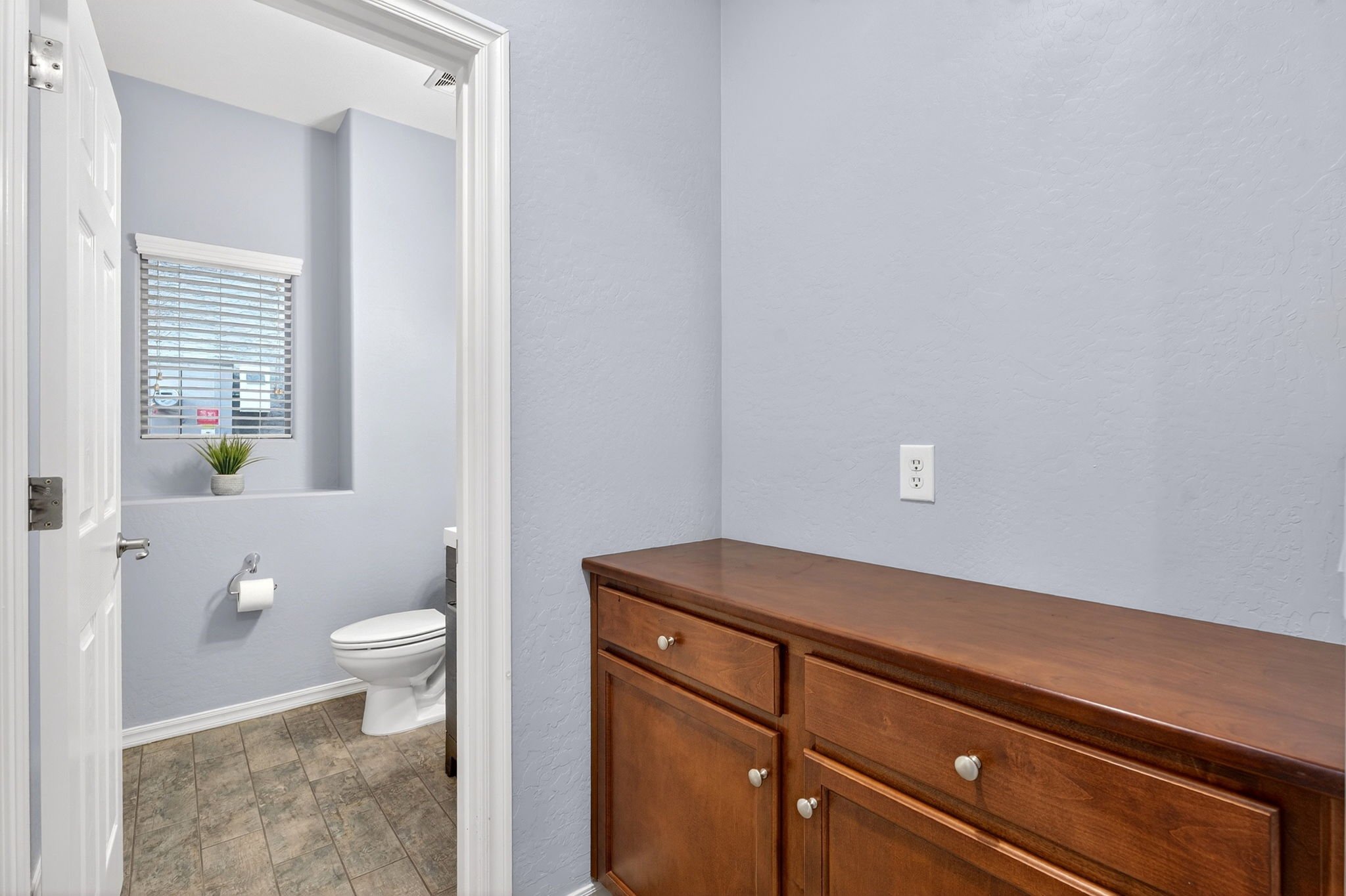 A small room with light gray painted walls, a wooden dresser, and a window with white blinds above the toilet in the bathroom which has tiled flooring.