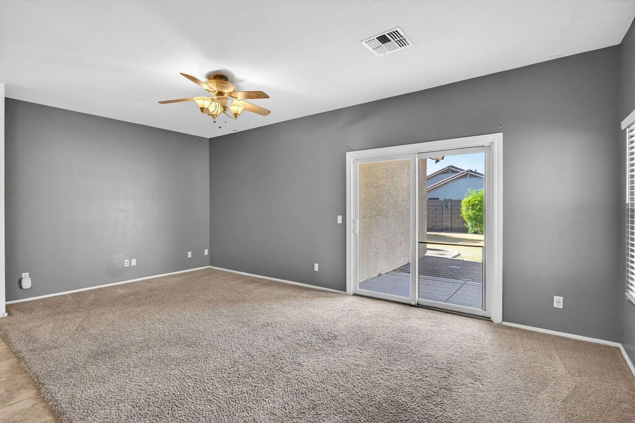 Empty living room with gray walls, beige carpet, sliding glass door leading outside, ceiling fan with lights.