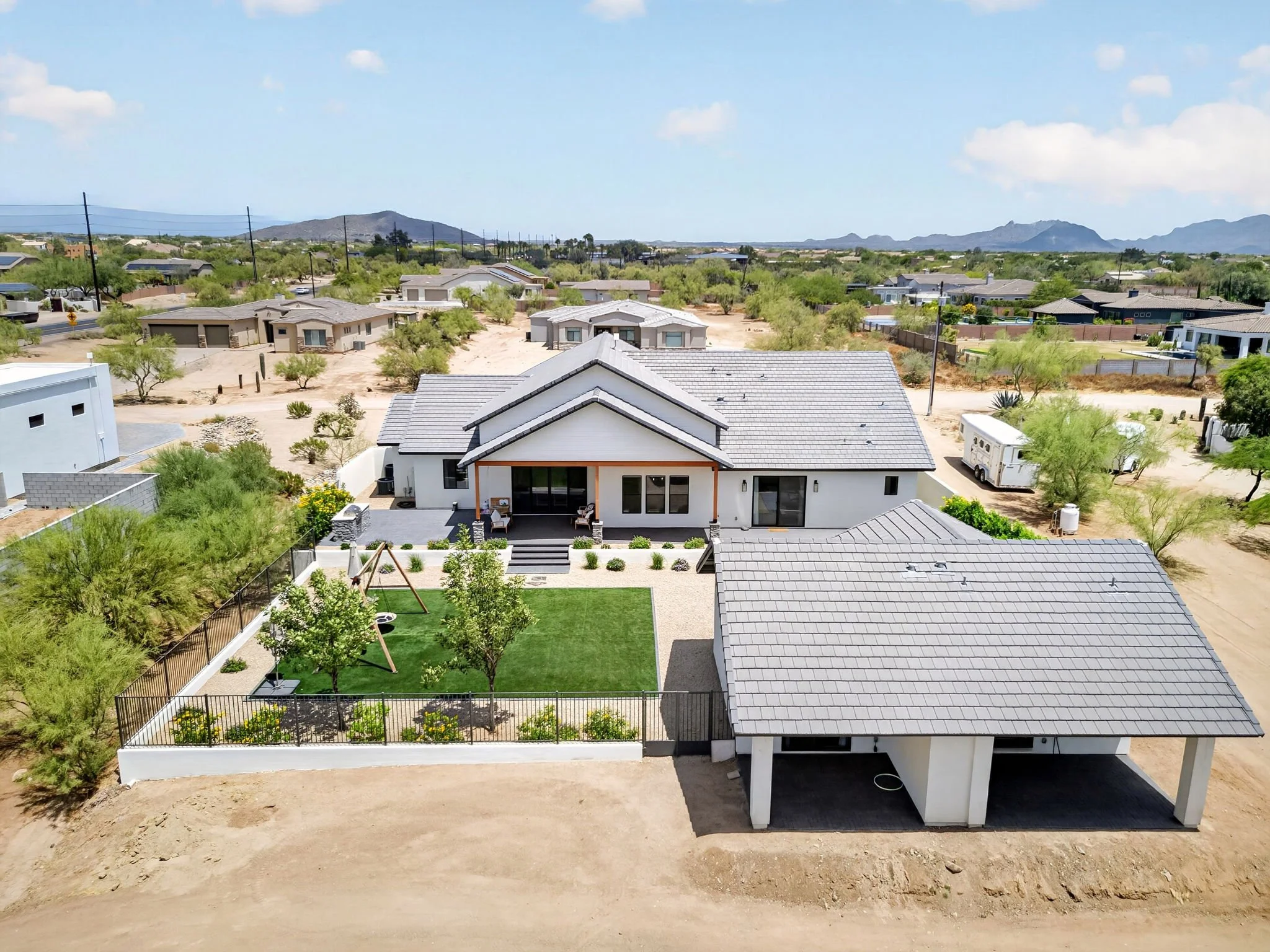 A residential backyard with a modern house, a green lawn, trees, a fence, and mountains in the background under a clear, partly cloudy sky.