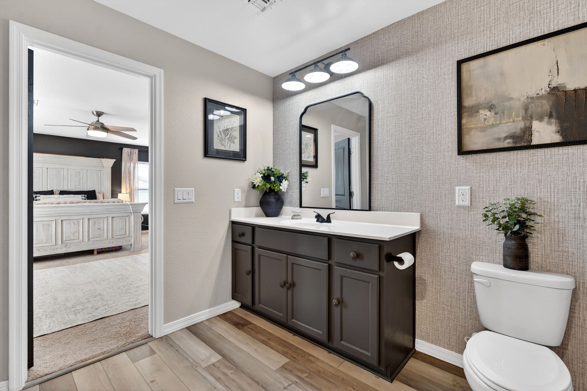 Bathroom with a dark vanity cabinet, a framed mirror, wall art, a flower vase, a toilet with a plant on top, and a textured wallpaper on one wall.
