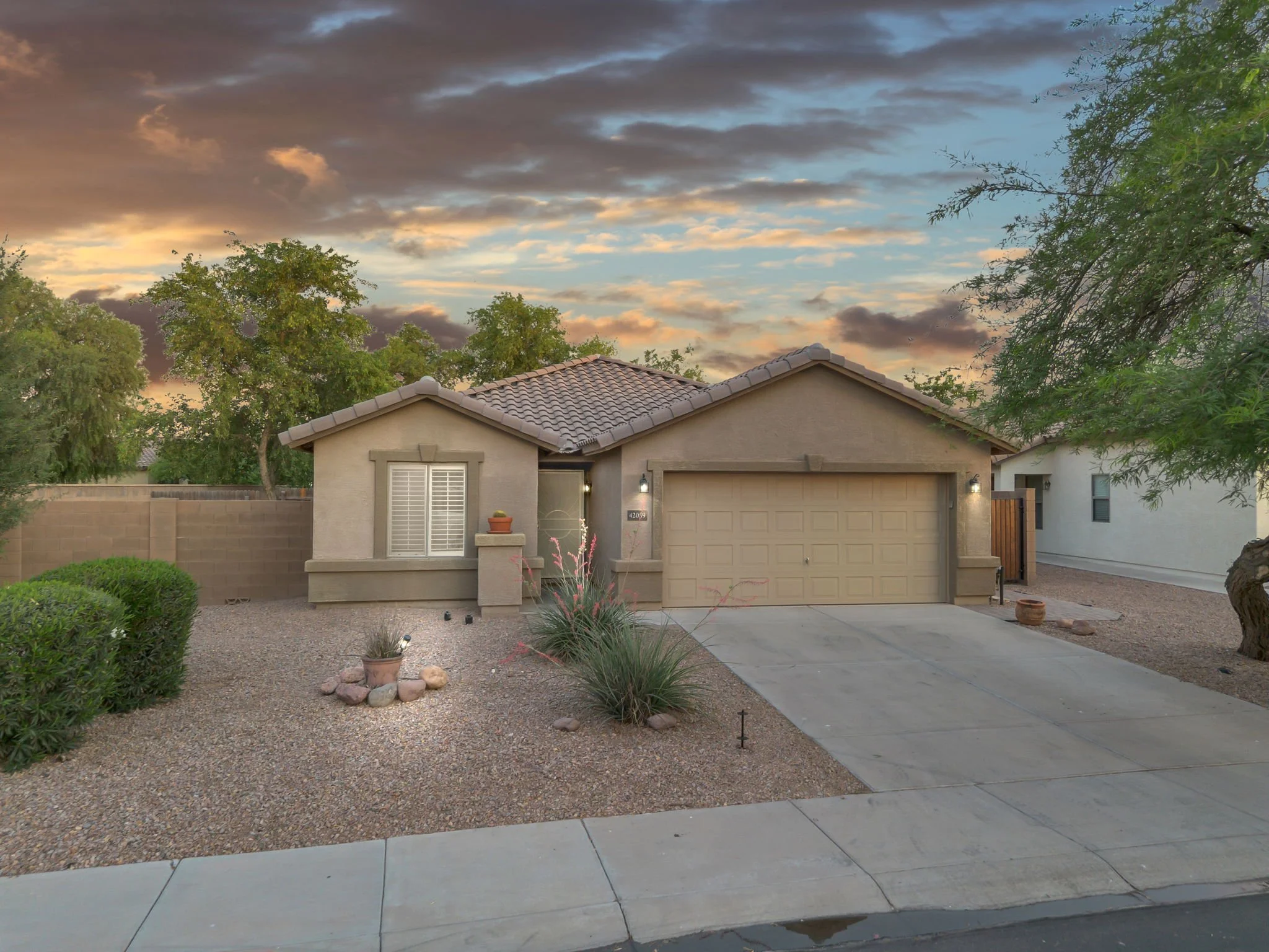 A suburban house with a beige exterior, front yard with gravel and desert plants, a large tree on the right, and a cloudy sky at sunset.