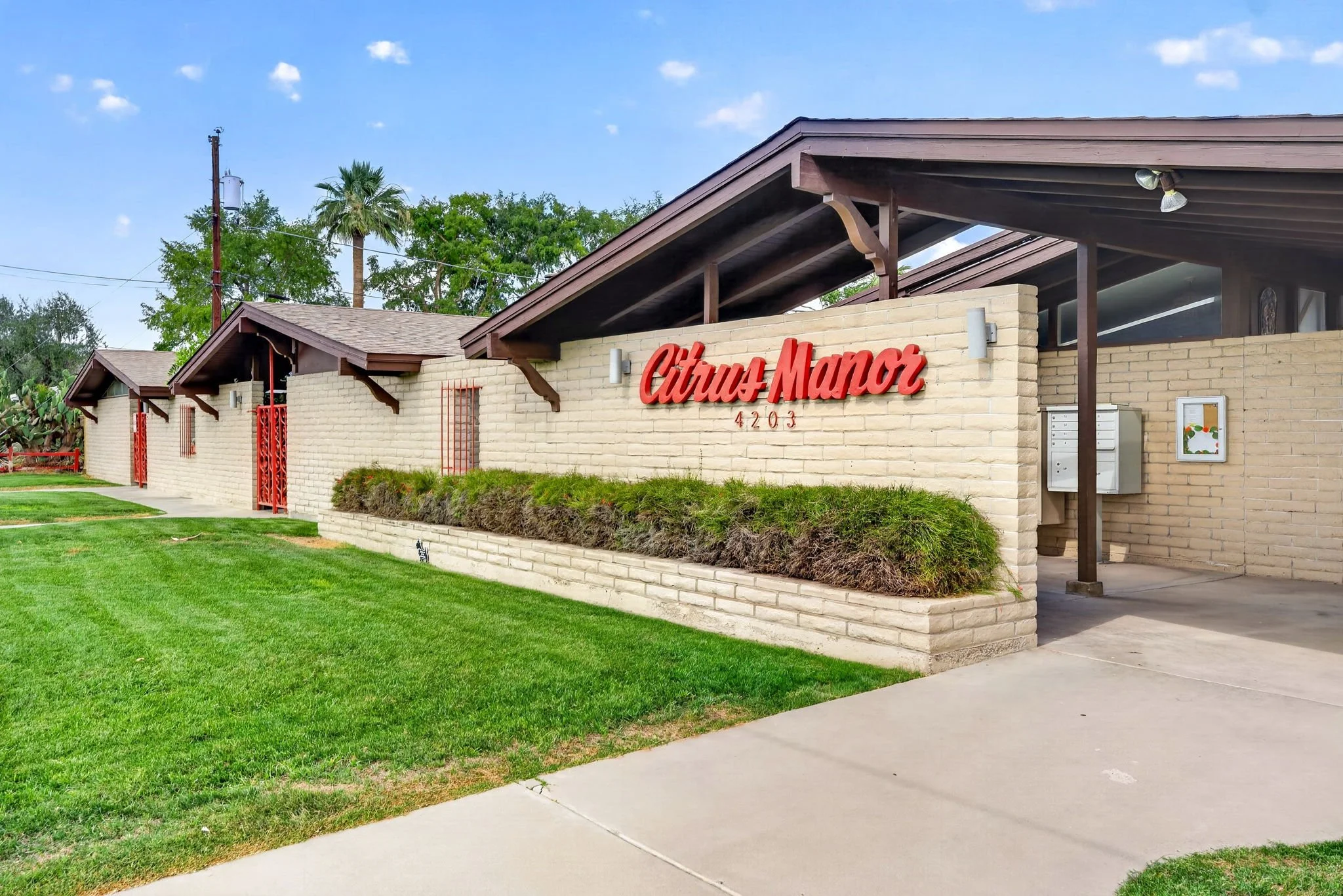 Exterior view of a modern residential building with a brick wall, brown roof, and red sign reading 'Citrus Manor' along with the number 4203. The foreground shows a well-maintained green lawn and a concrete sidewalk, under a partly cloudy sky.