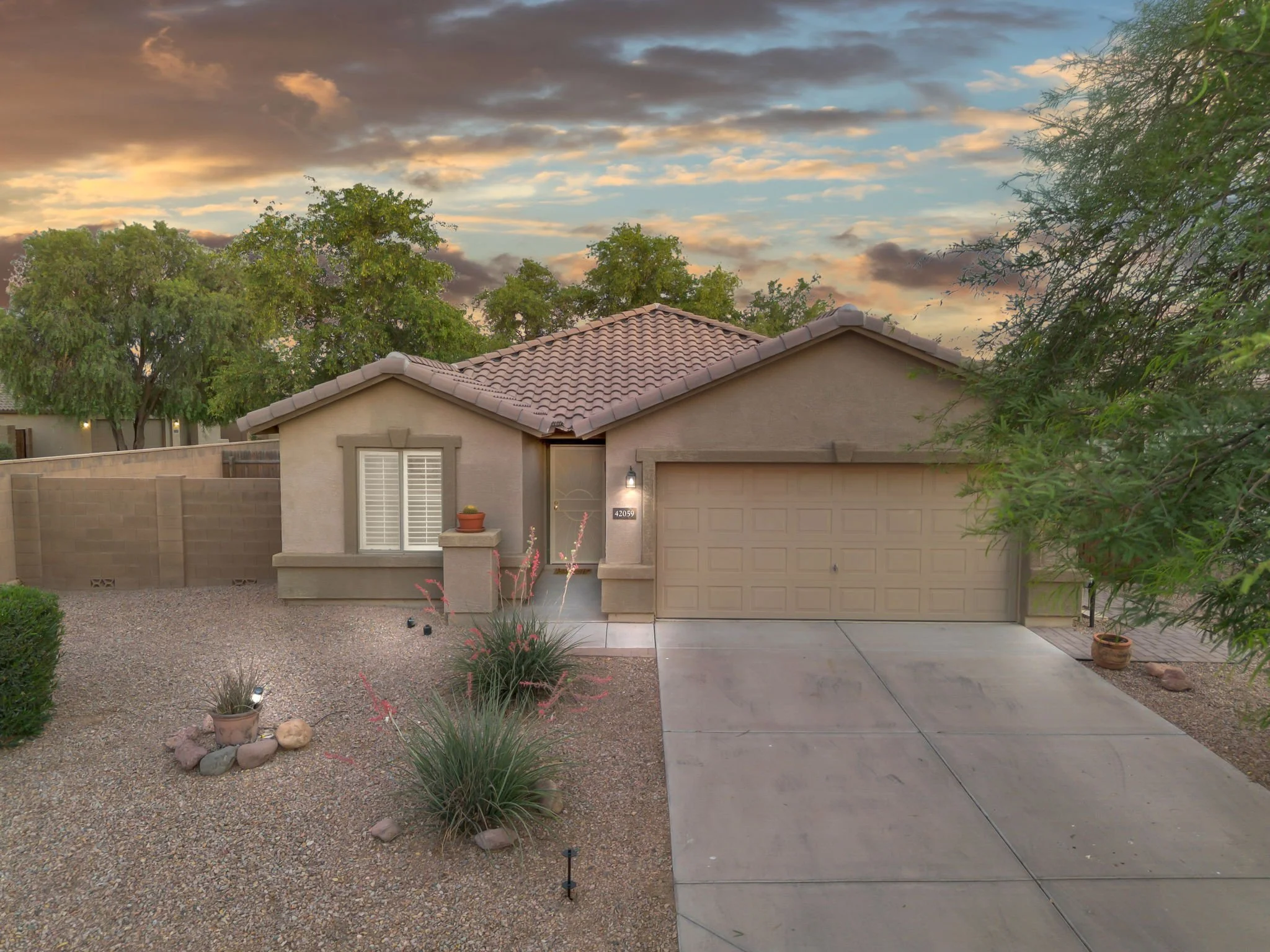 Front yard with desert landscaping, small rocks, plants, and a concrete pathway leading to a beige house with a tiled roof and a garage, under a partly cloudy sky at sunset.