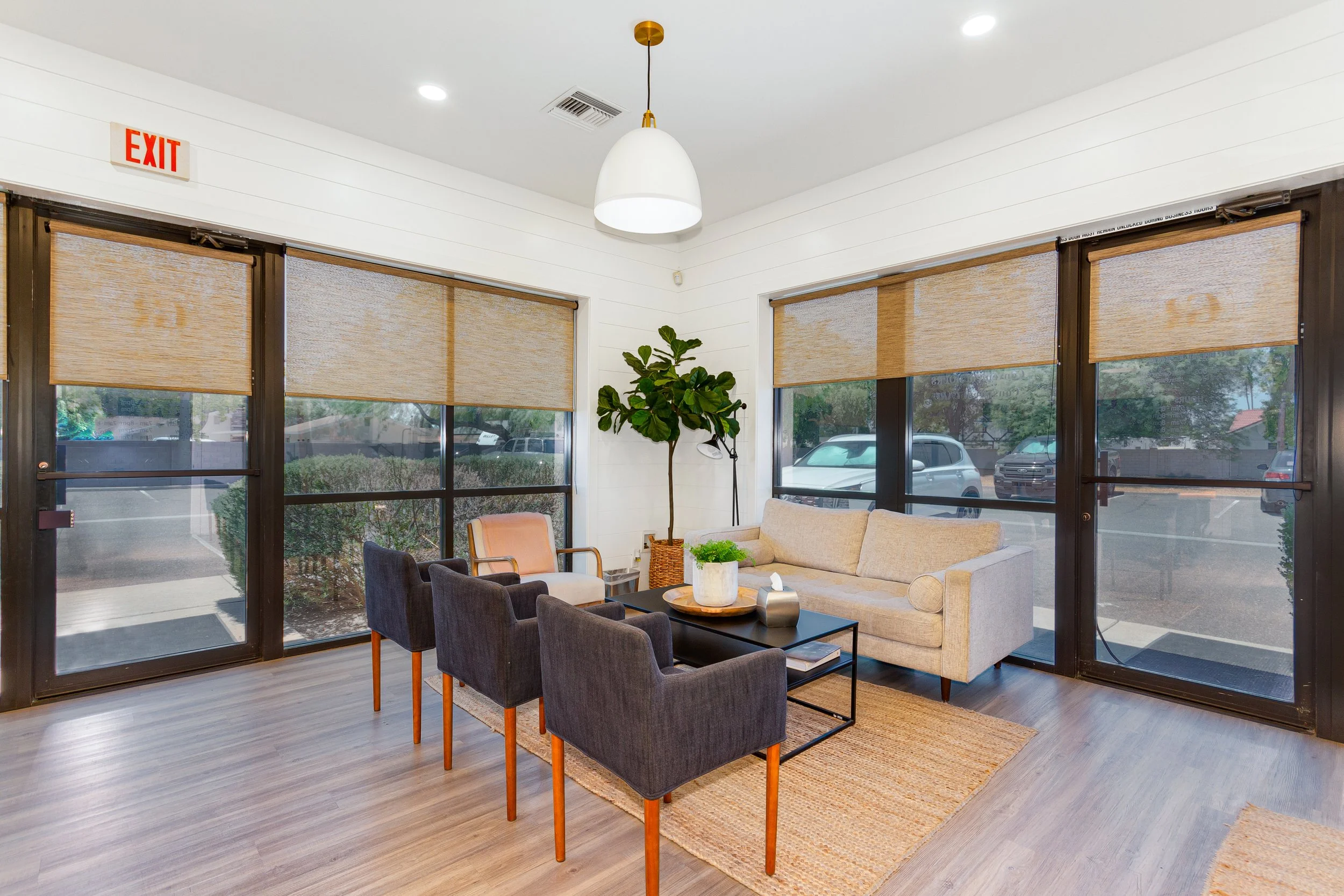 Interior of a bright, cozy waiting or lounge area with large windows covered with beige blinds. There are several chairs, a beige sofa, a black coffee table with books and a decorative tray, a potted plant, and a white pendant light hanging from the 