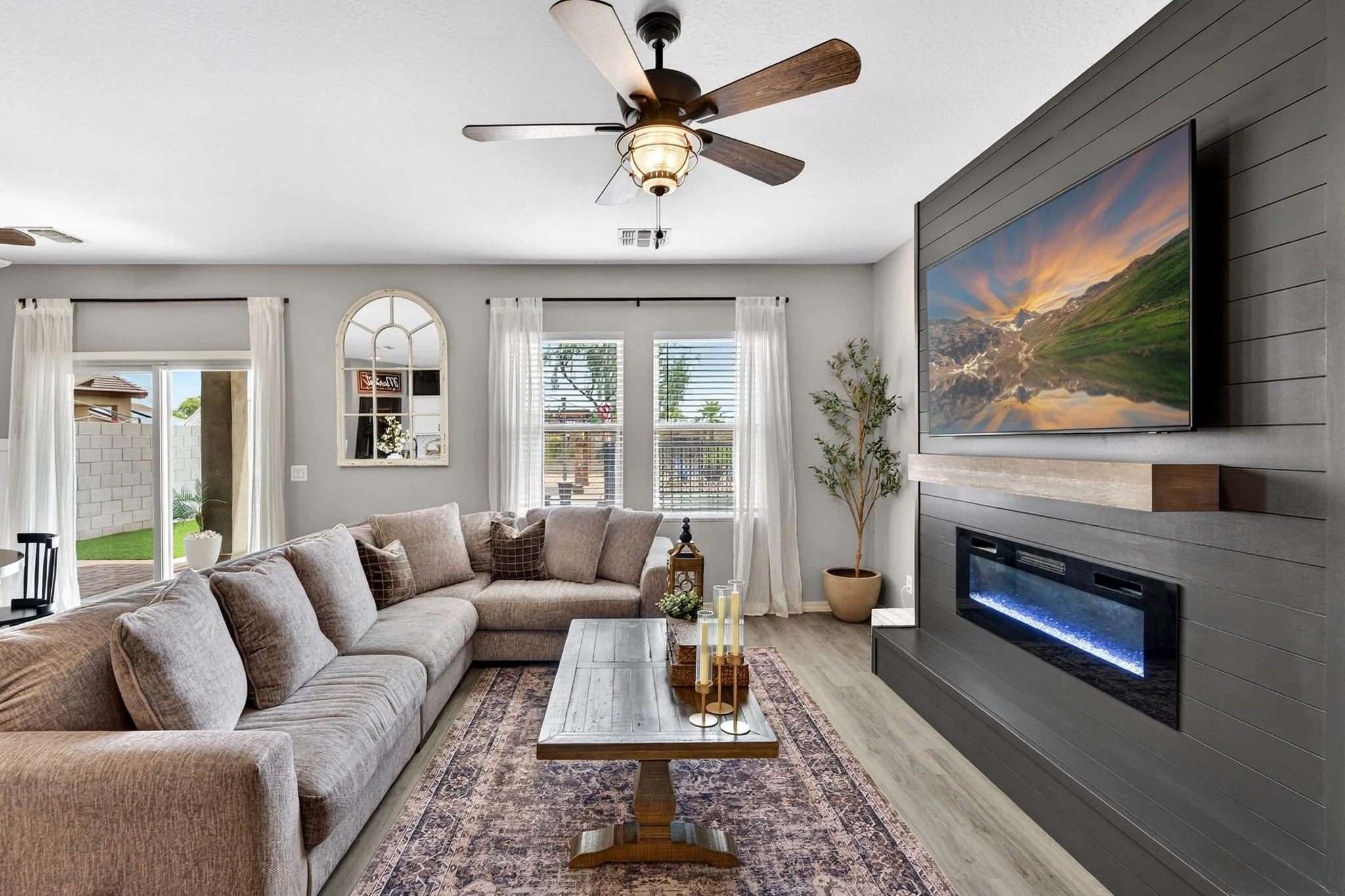 Living room with a beige sectional sofa, wooden coffee table, fireplace with a landscape television, large windows with white curtains, ceiling fans, and a potted plant.