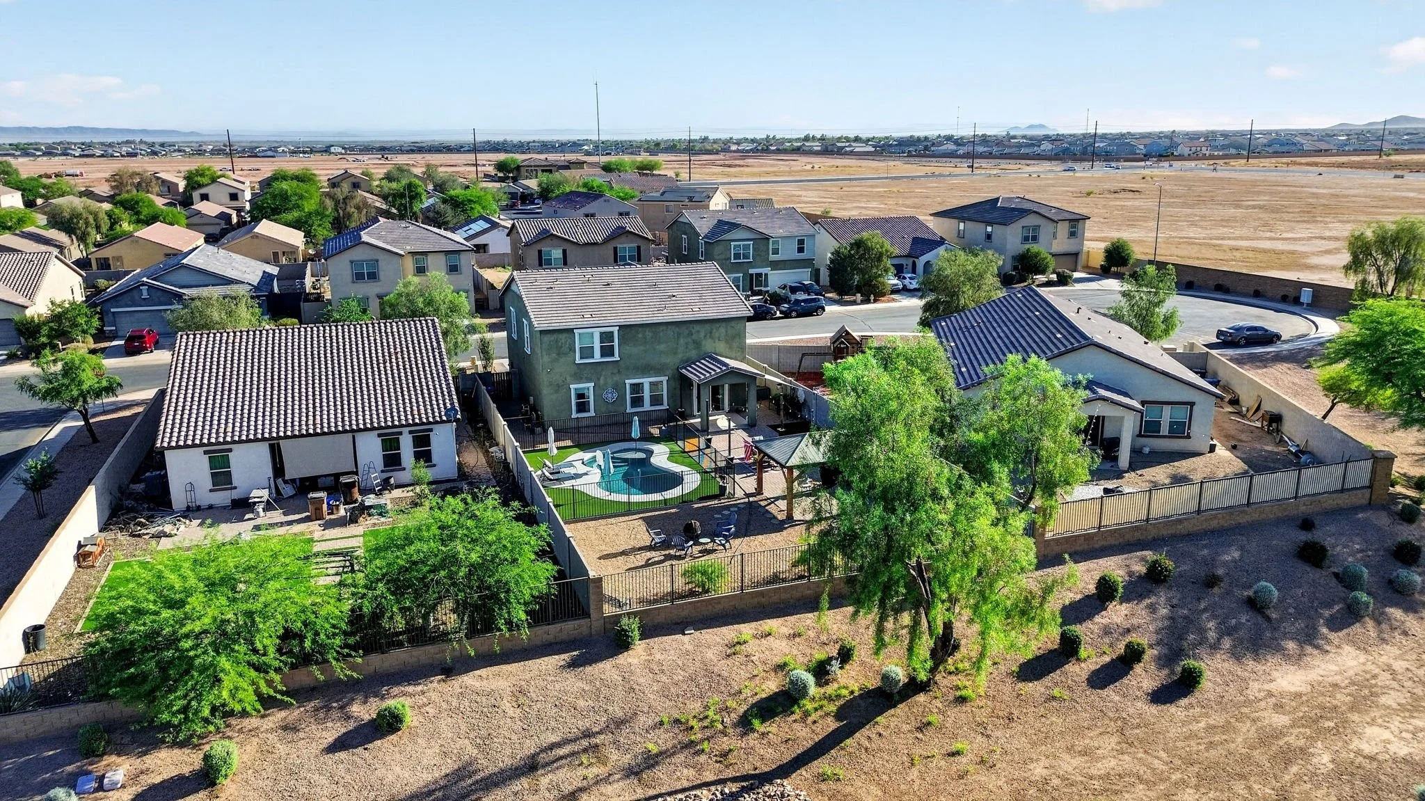 A view of a suburban neighborhood with houses, trees, and a backyard pool, with a barren field and distant mountains in the background.