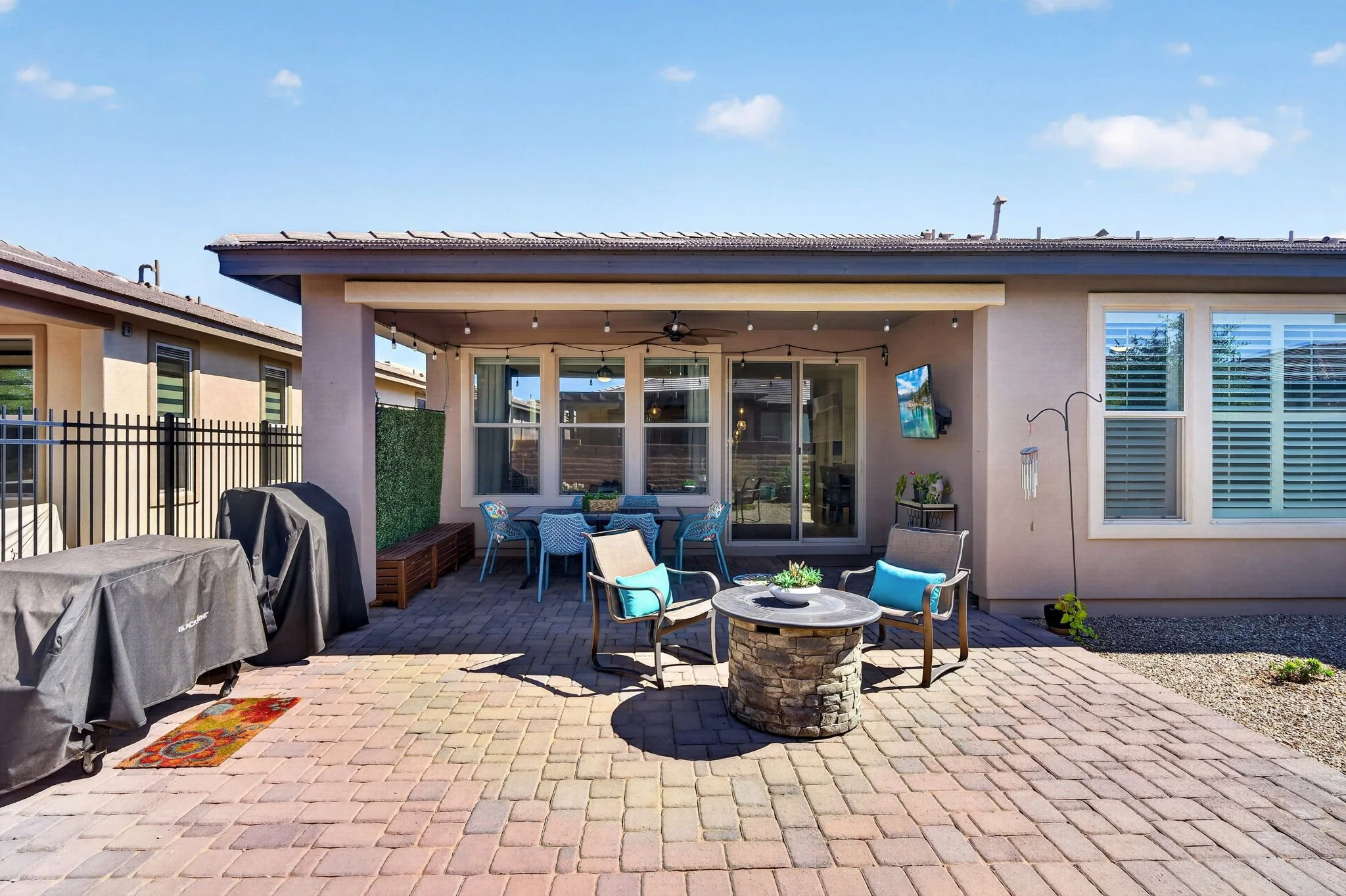 Backyard patio with brick pavers, outdoor seating, a grill covered with a gray cover, string lights, and a wall-mounted TV, with a house featuring large windows in the background.