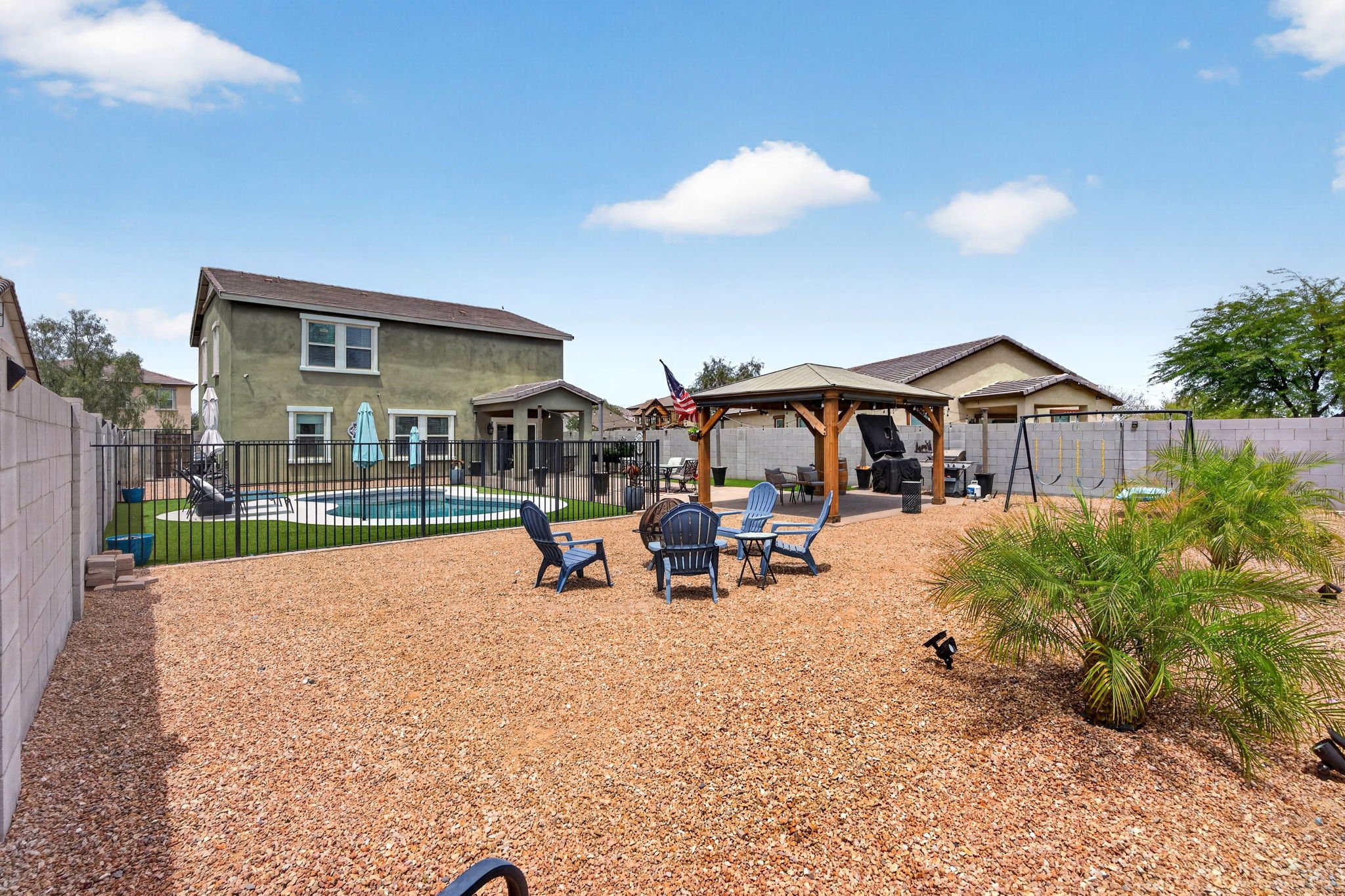 Backyard with swimming pool, outdoor table and chairs, covered patio, and lawn chairs, enclosed by cinder block wall, with two houses and trees under a partly cloudy sky.