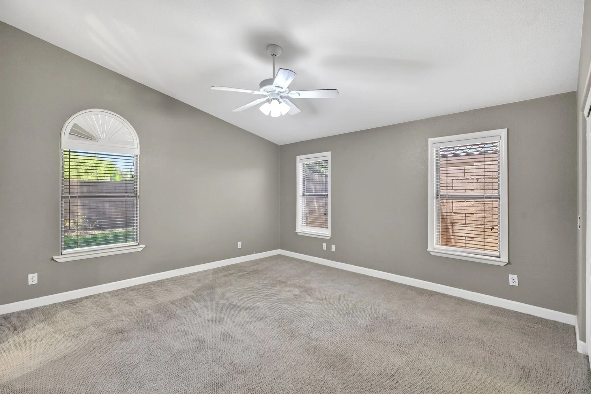 Empty living room with gray walls, three windows with white blinds, a white ceiling fan, and beige carpet.