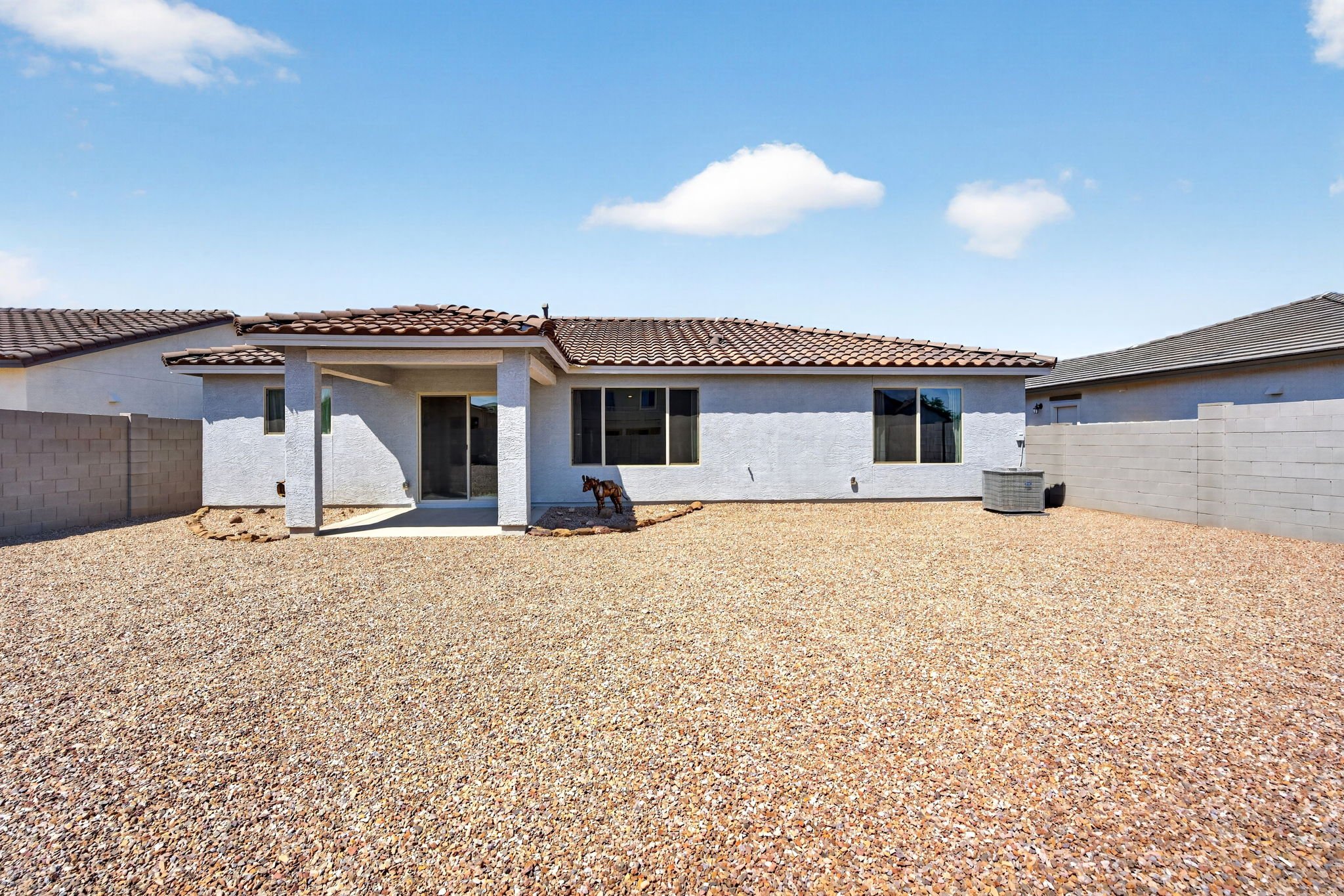 Backyard with gravel ground, white house with red tiled roof, sliding glass door, windows, residence next door, small dog statue, air conditioning unit, and brick wall fencing under a partly cloudy sky.