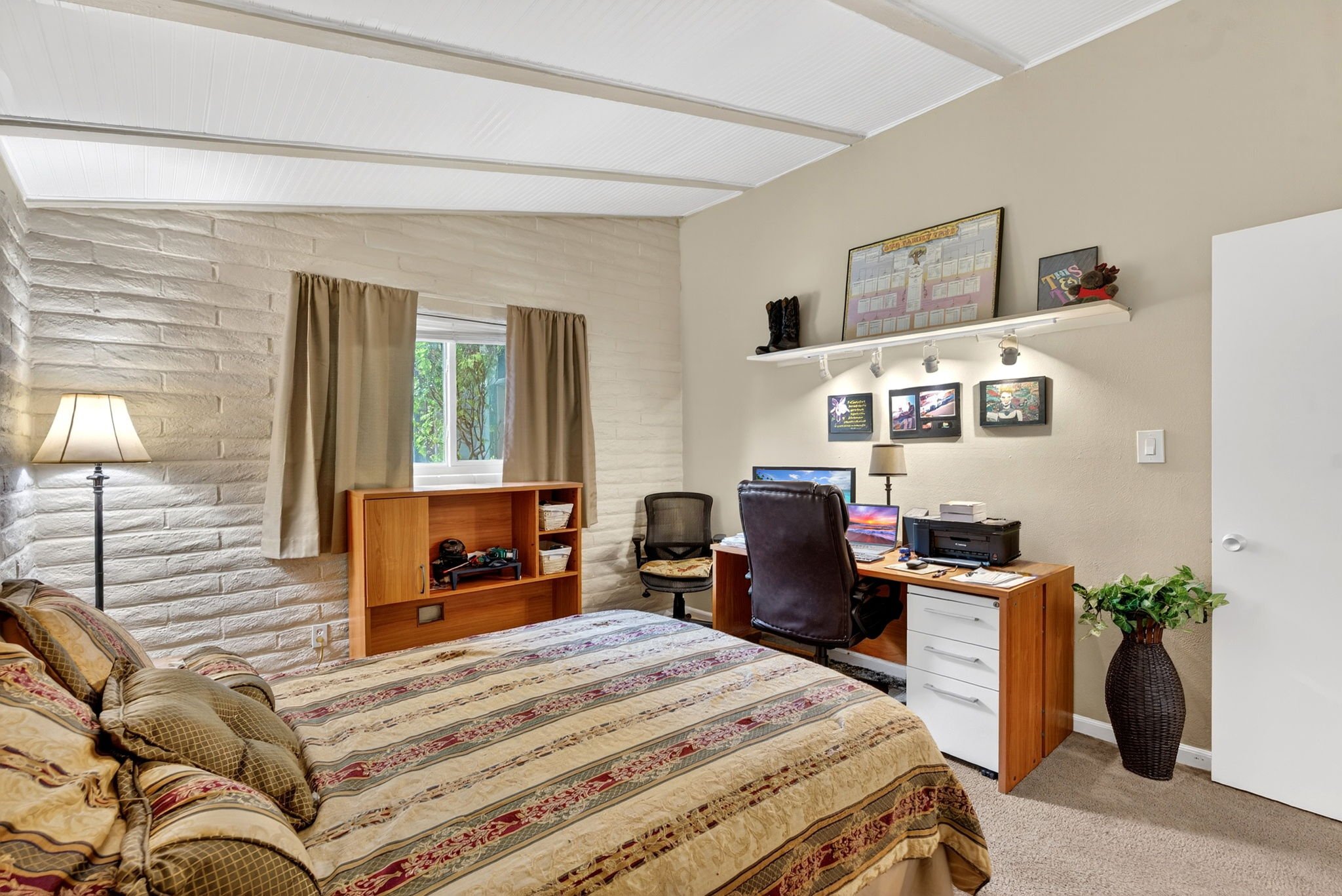 A cozy bedroom with a large bed with a patterned bedspread and multiple pillows, a wooden bookshelf, a seated office chair, a desk with a computer, and wall decor including framed pictures and a shelf with plush toys and decorations. A window with be
