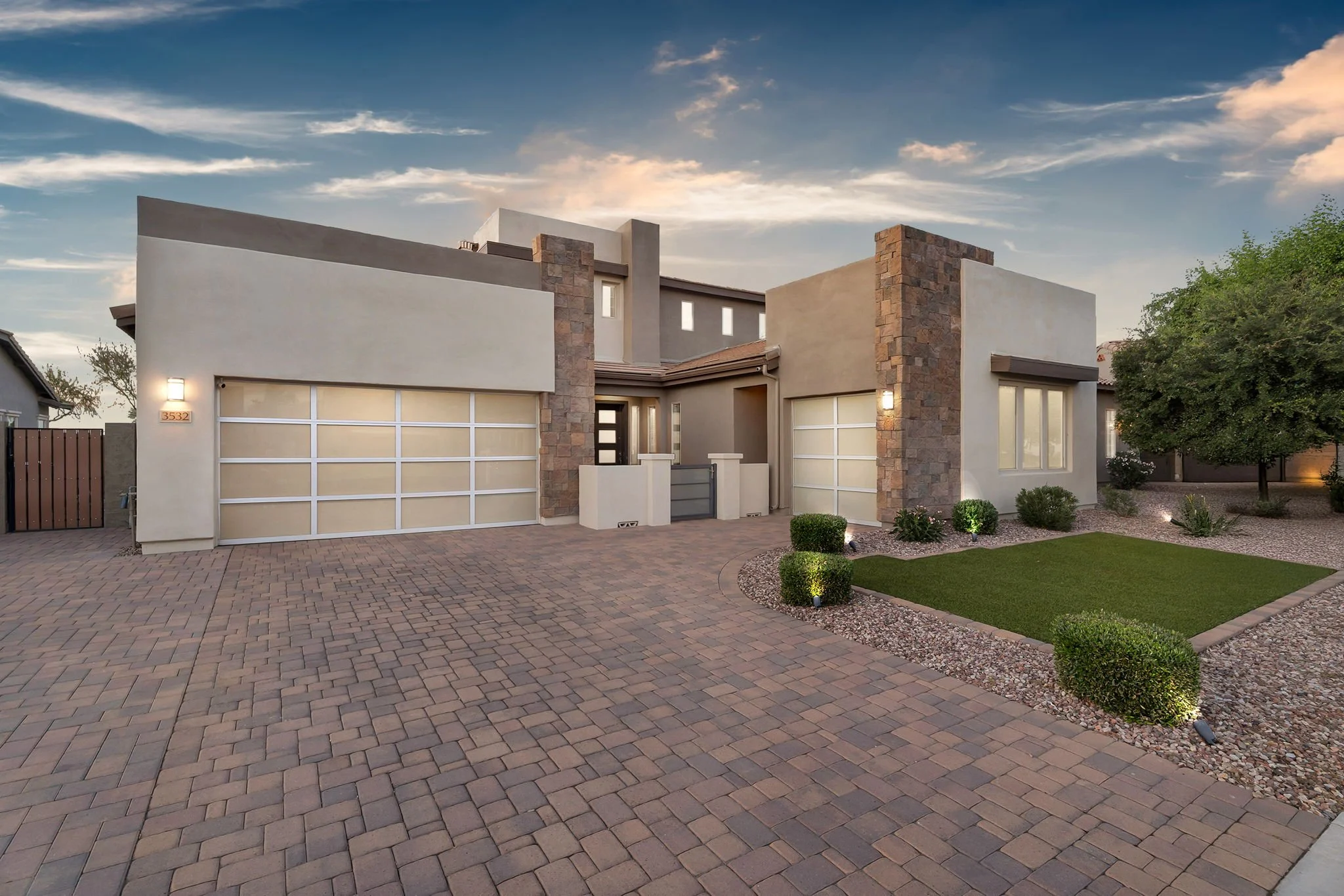 Modern house with a beige facade, stone accents, and a white garage door, surrounded by a landscaped yard with bushes, a small lawn, and brick pavement, under a partly cloudy sky.