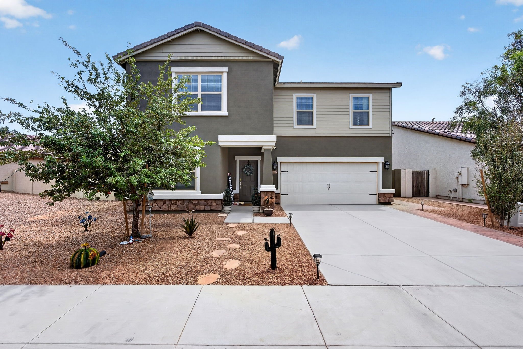 Front view of a two-story modern house with a gray and beige exterior, white-framed windows, a garage door, and a small front yard decorated with cacti and desert plants, under a blue sky with some clouds.