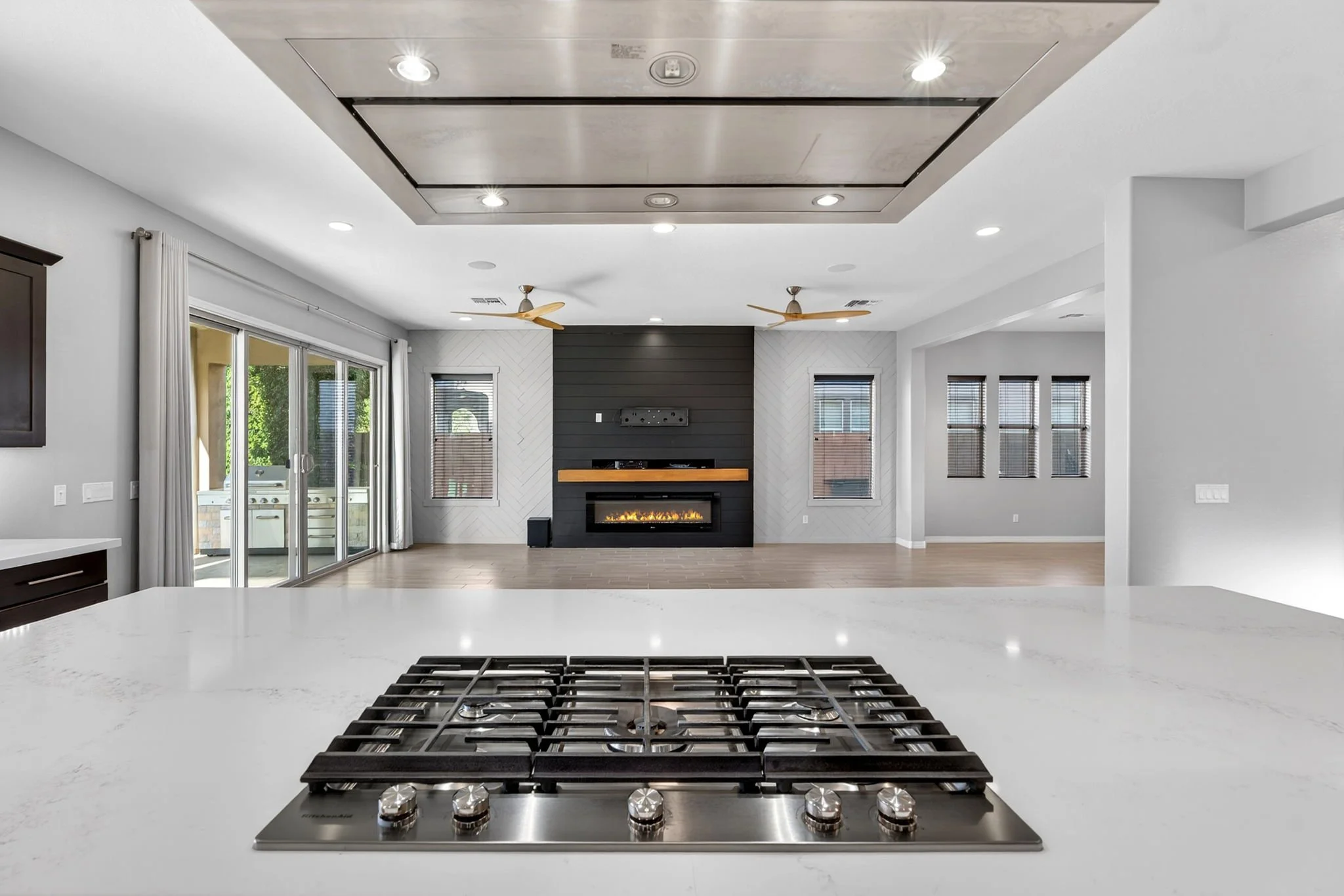 Modern kitchen with a white countertop in the foreground, a black gas stove, and a living room with windows, a fireplace, and ceiling fans in the background.