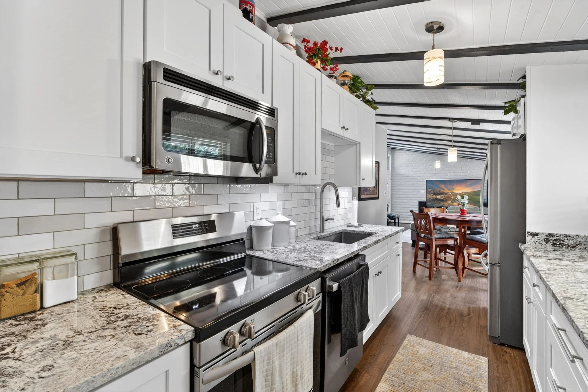 A modern kitchen with white cabinets, granite countertops, a stainless steel oven and microwave, and a white tiled backsplash. There is a sink with a faucet, some jars, and a window with a view of the dining area and living room with a television and
