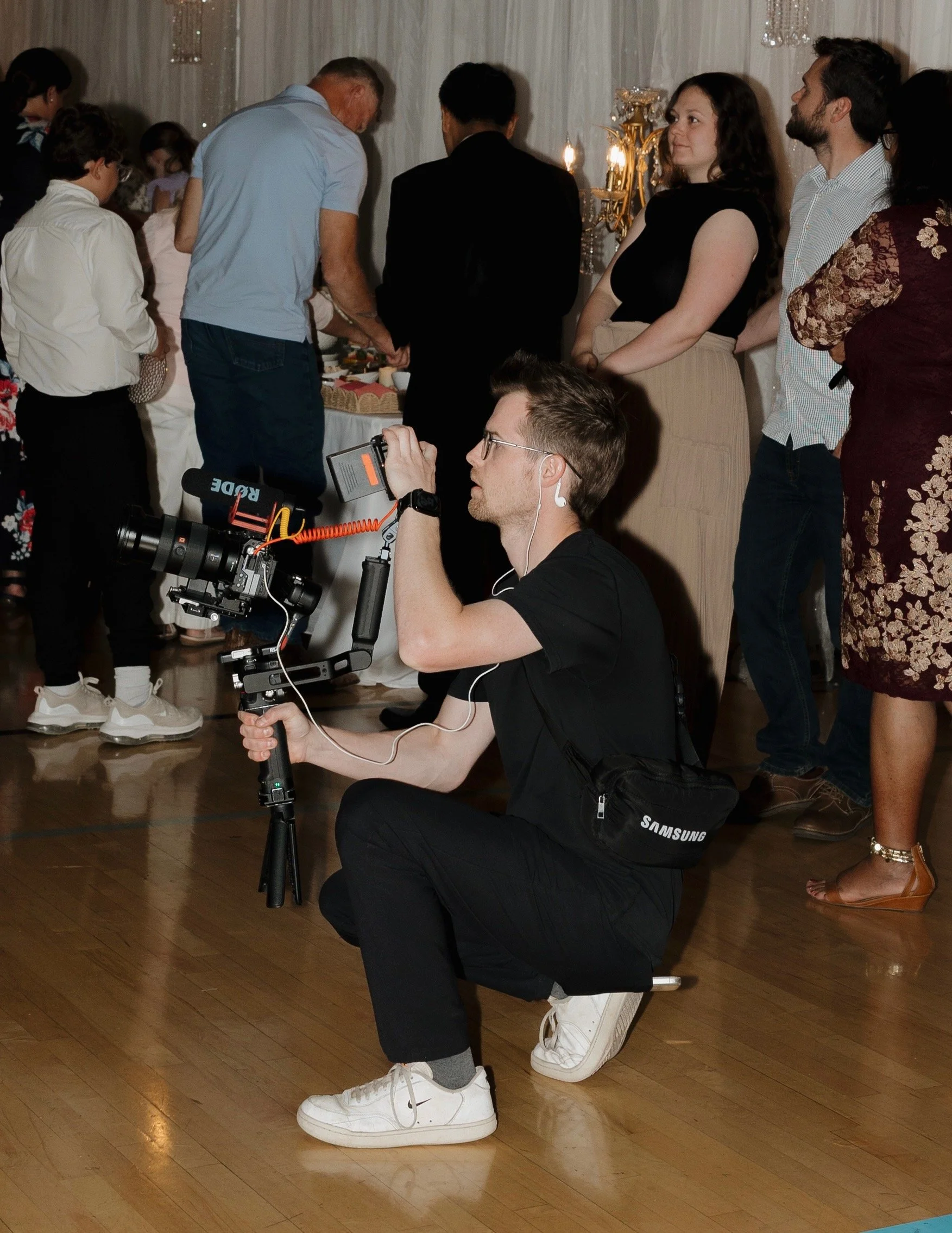 A group of people at an indoor event, some standing and one person kneeling with a camera on a stabilizer, capturing footage. The setting appears to be a celebration or gathering.