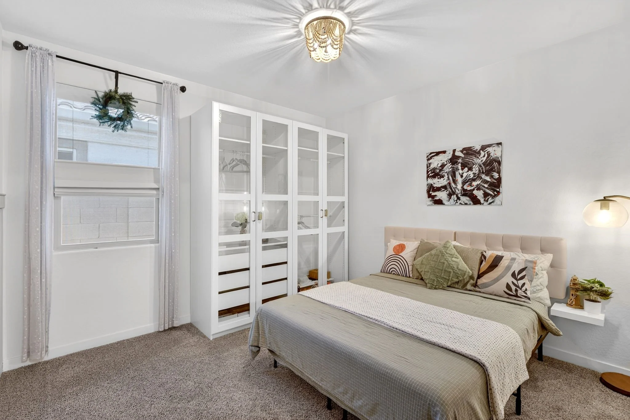 A bedroom featuring a bed with beige and patterned pillows, a white nightstand with decorative items, a white wardrobe with glass doors, a small window with sheer curtains and a small wreath, artwork on the wall, and a ceiling light fixture.