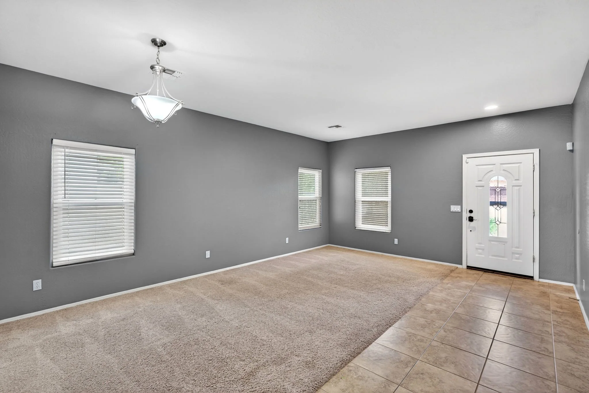 Living room with gray painted walls, three windows with blinds, tiled and carpeted flooring, a ceiling light fixture, and a front door with a glass panel.