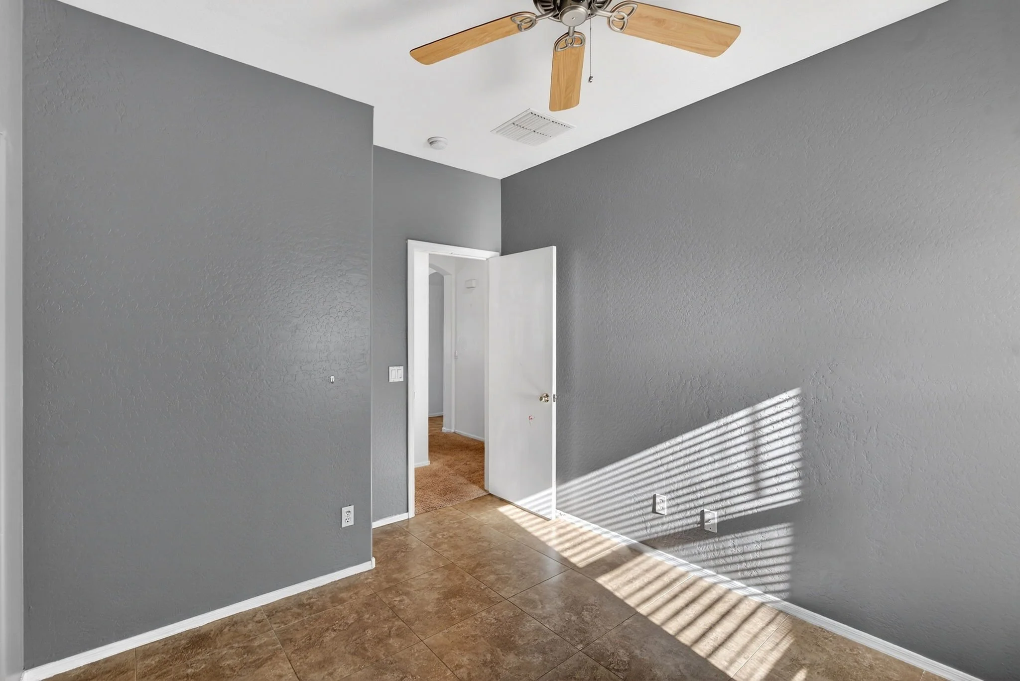 Empty room with gray painted walls, brown tiled flooring, an open door leading to another room with ladder-like carpet, a ceiling fan with wooden blades, and sunlight creating shadow stripes on the wall.